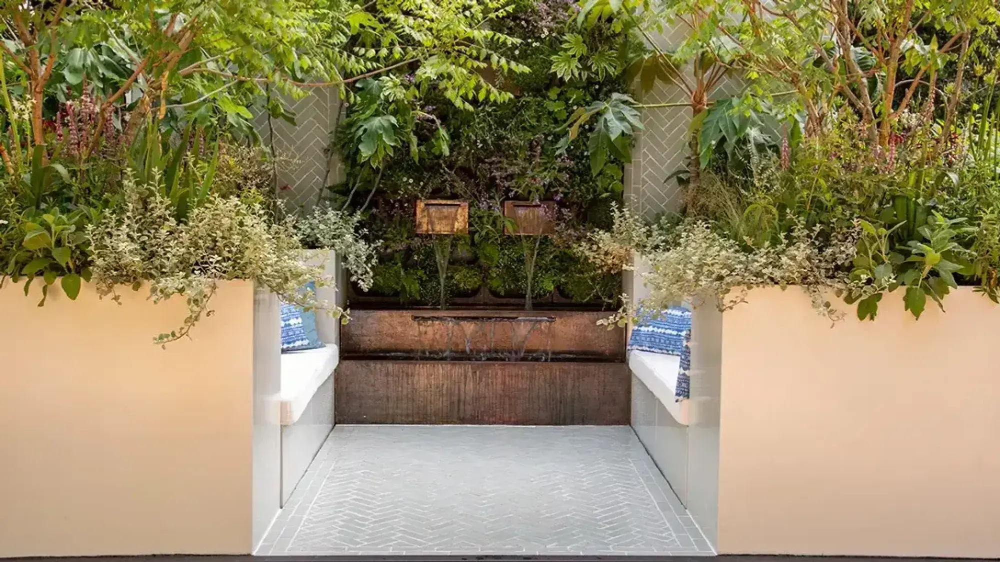 A lush, modern container courtyard featuring large cream planters, built-in benches, and a multi-tier copper-finish water feature set into a vibrant green living wall.