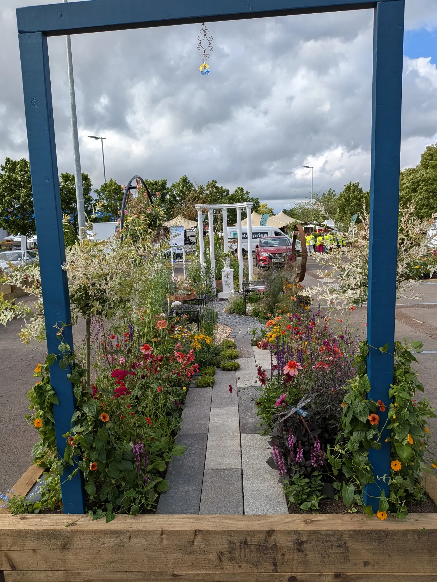 A blue timber archway frames a vibrant garden border with a curving paved path, flanked by colourful dahlias, salvias, and variegated willow trees.