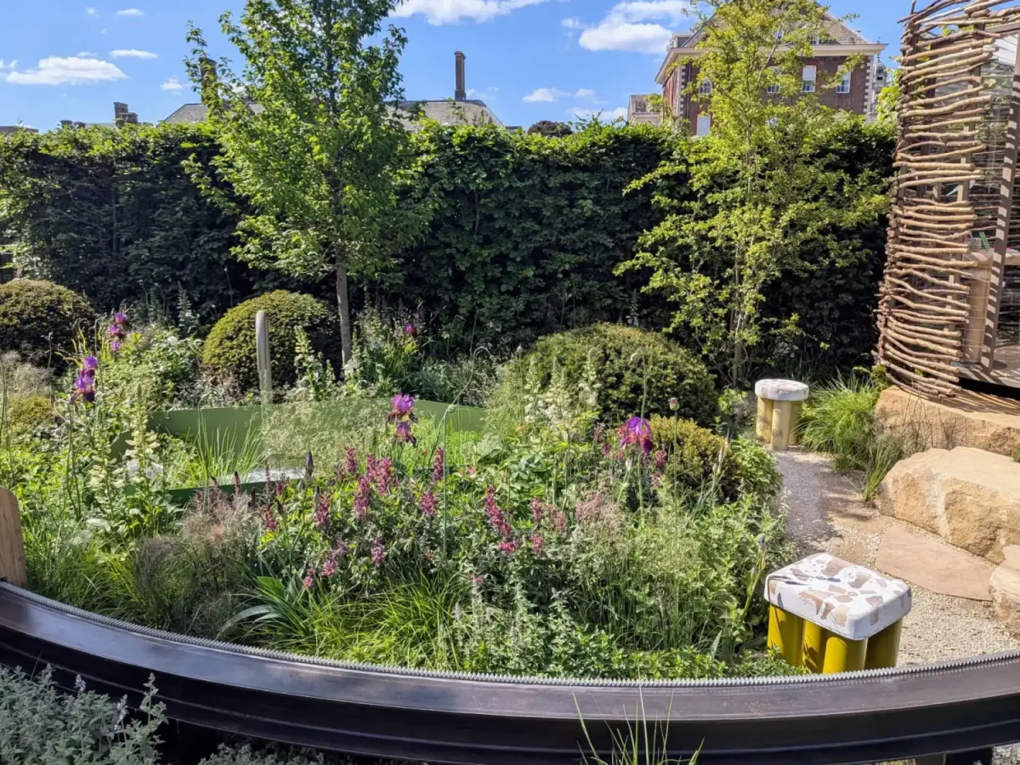 A naturalistic garden with a gravel path, woven wood den, yellow mosaic stools, and lush planting of purple and pink flowers.