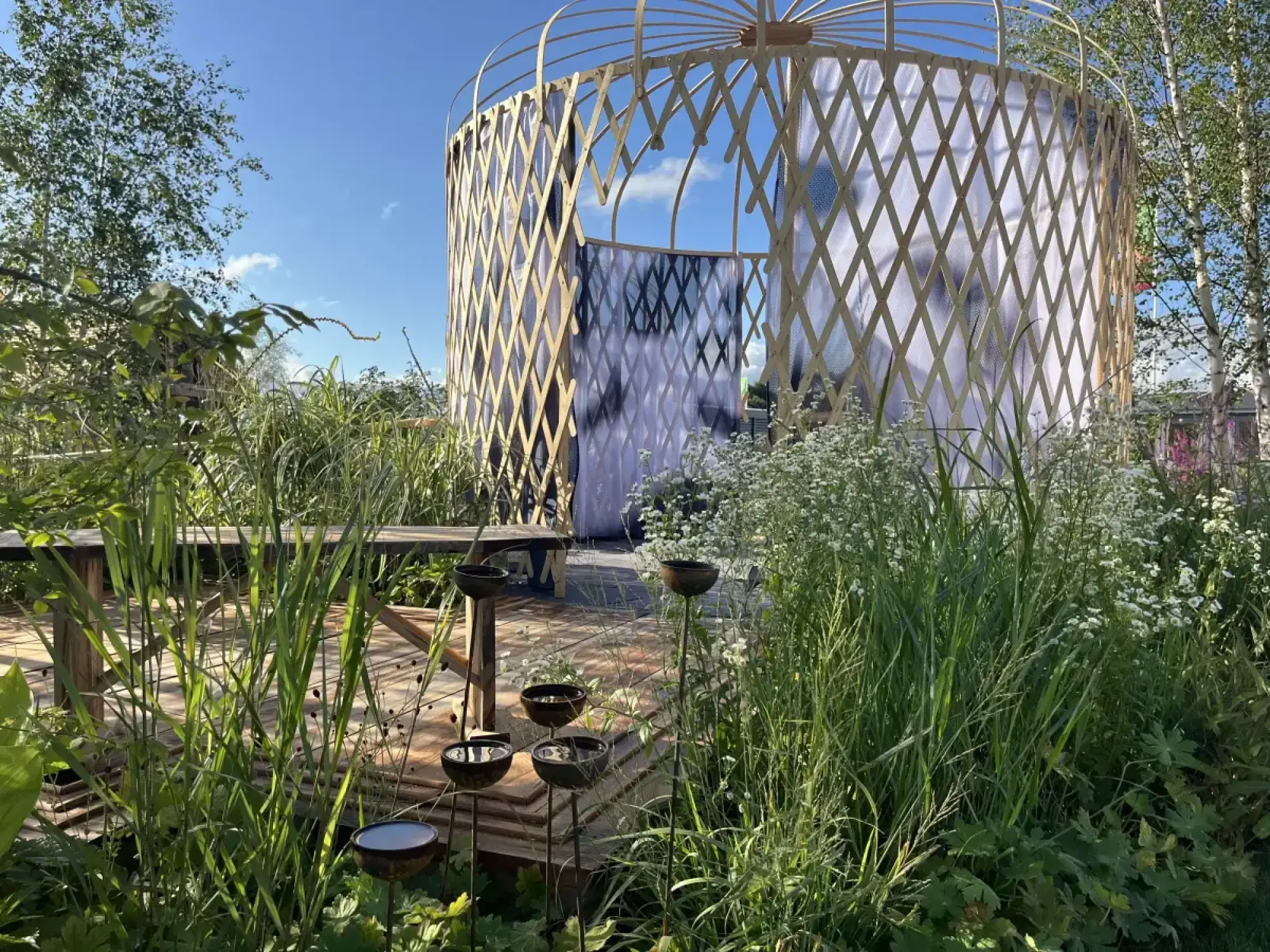 A circular structure with white panels overlooks a wooden deck, lush grasses, flowers, and water bowls.
