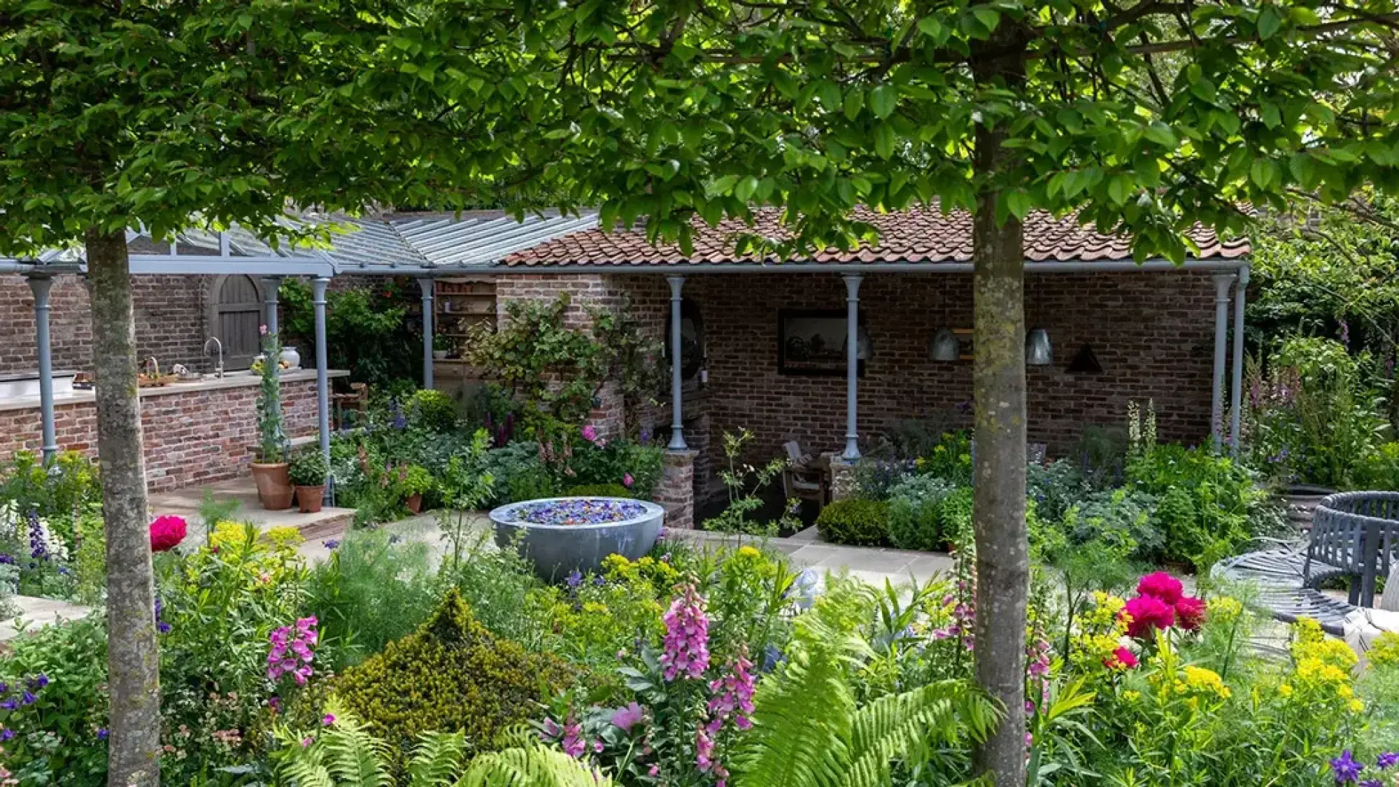 A lush show garden features a covered outdoor kitchen and a brick outbuilding with a terracotta roof, surrounded by vibrant pink, yellow, and purple flowers and a central zinc water bowl.