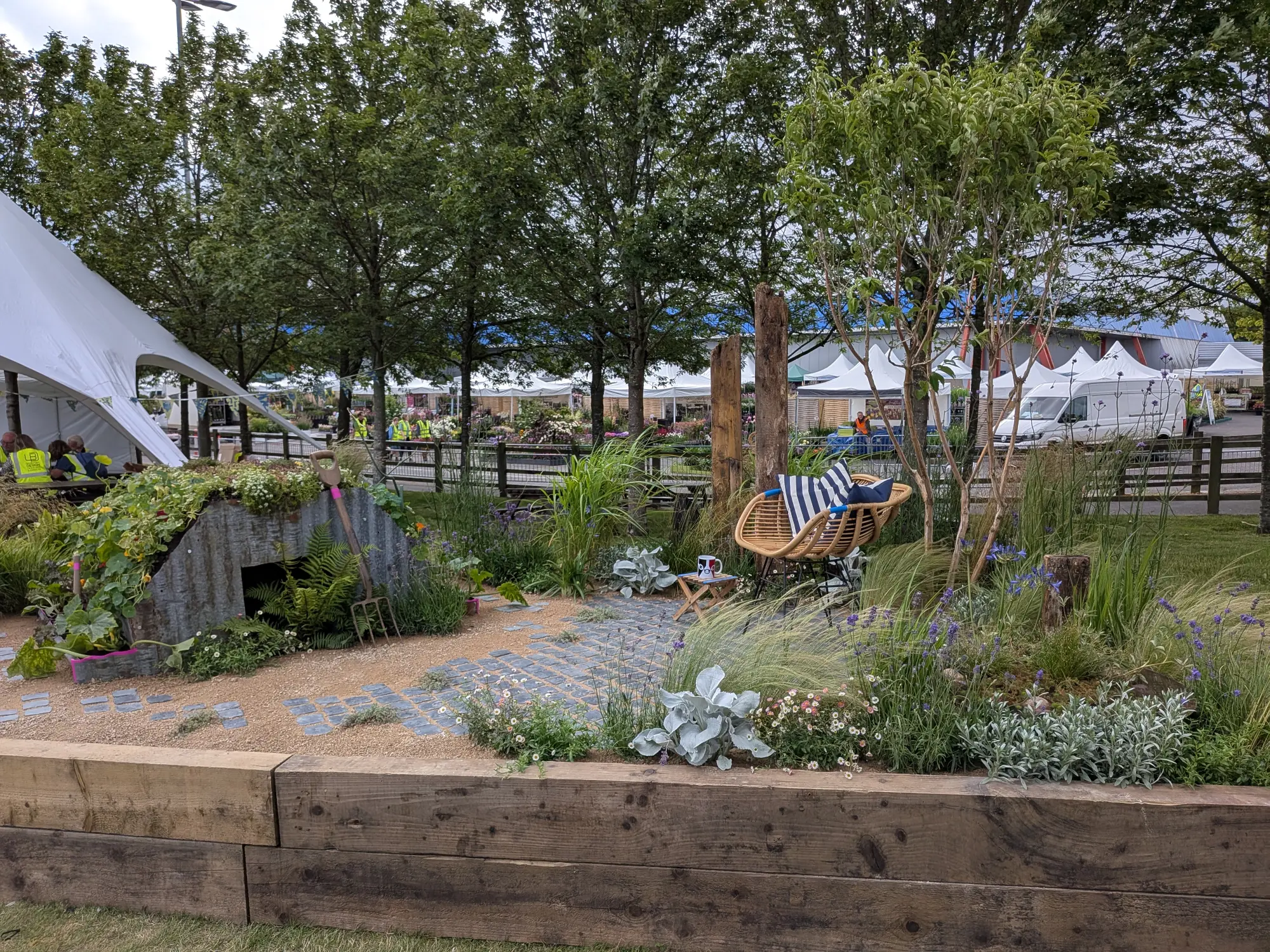 A beautiful border garden featuring a corrugated iron shelter covered in climbing plants, a gravel path with slate pavers, a rattan chair, and naturalistic planting with grasses, silvery foliage, and purple flowers.
