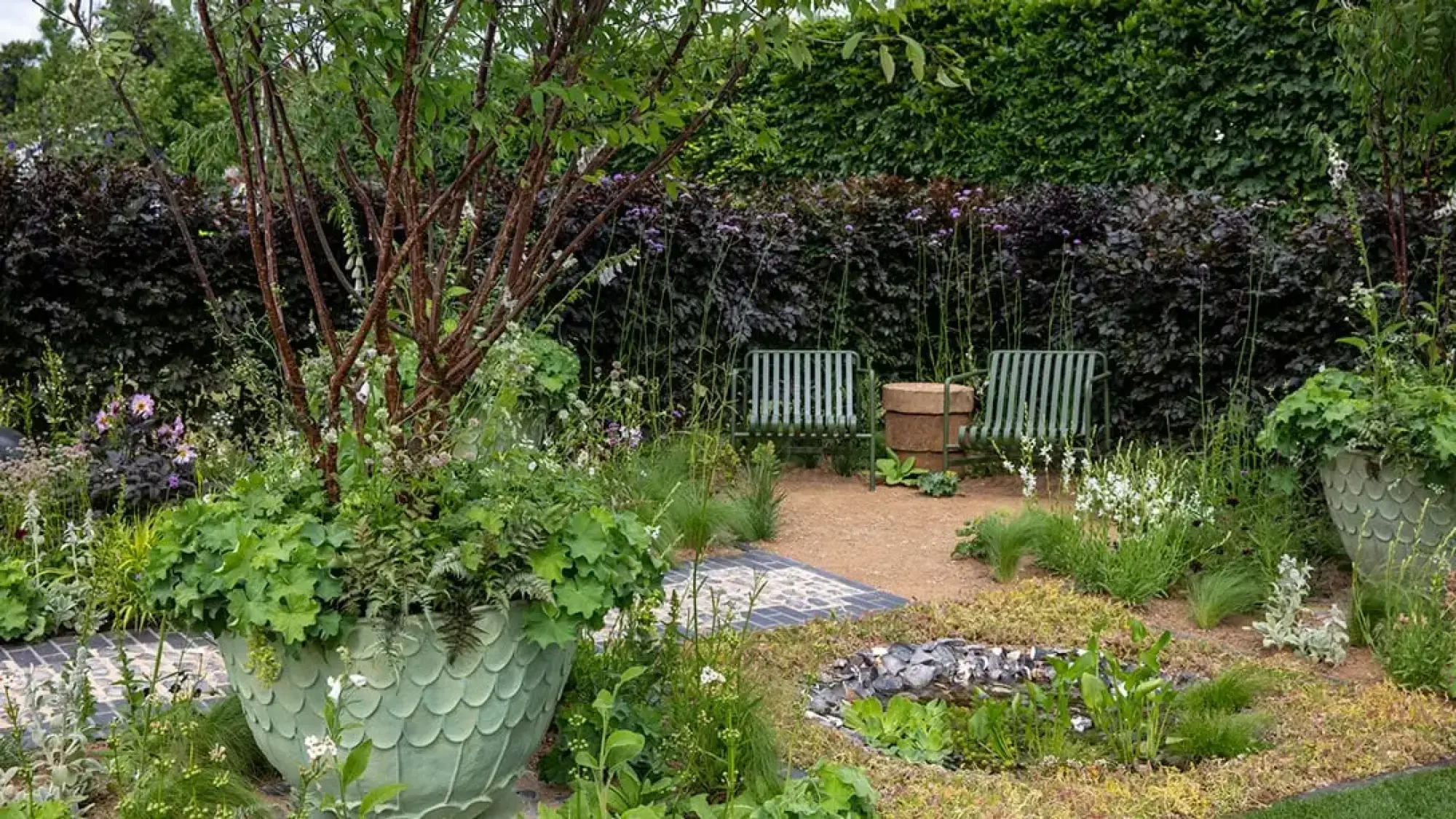 A tranquil traditional townhouse garden features a large green scaly planter, an informal pond, and green slatted chairs among lush mixed planting and dark hedges.