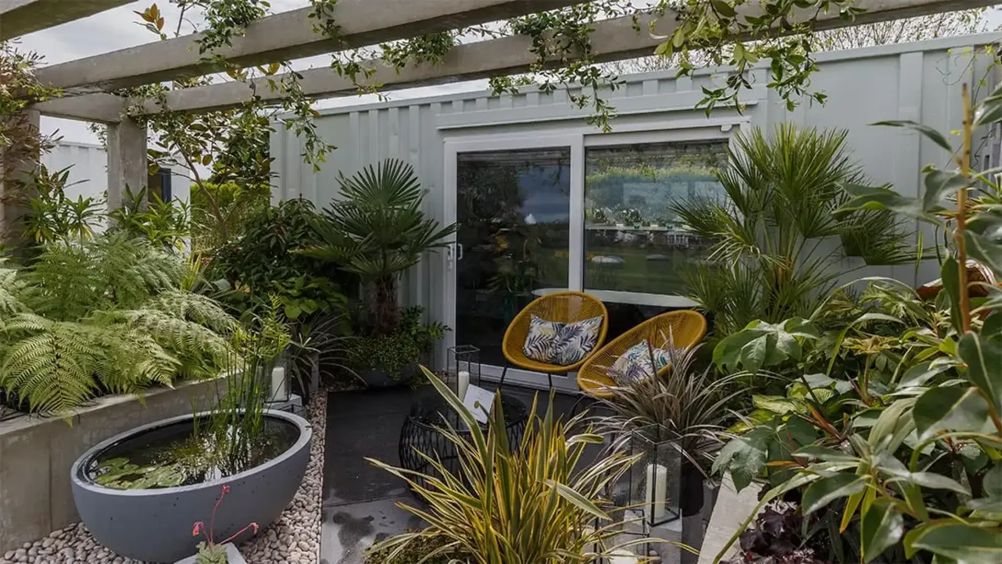 A green living space features two yellow woven chairs, ferns, palms, an oval pond planter, and a concrete pergola.