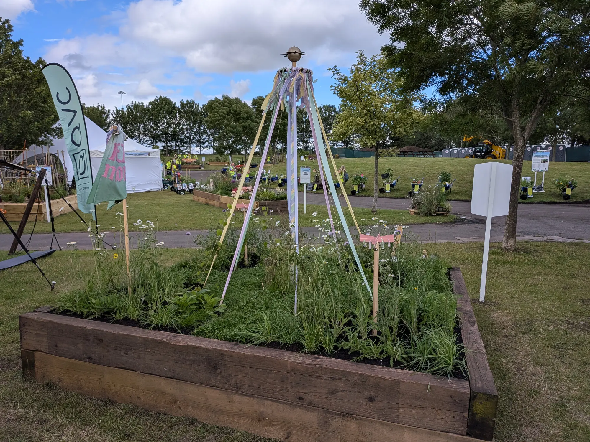A raised timber border garden features a central maypole with pastel ribbons, surrounded by informal planting of white daisy-like flowers and green foliage.