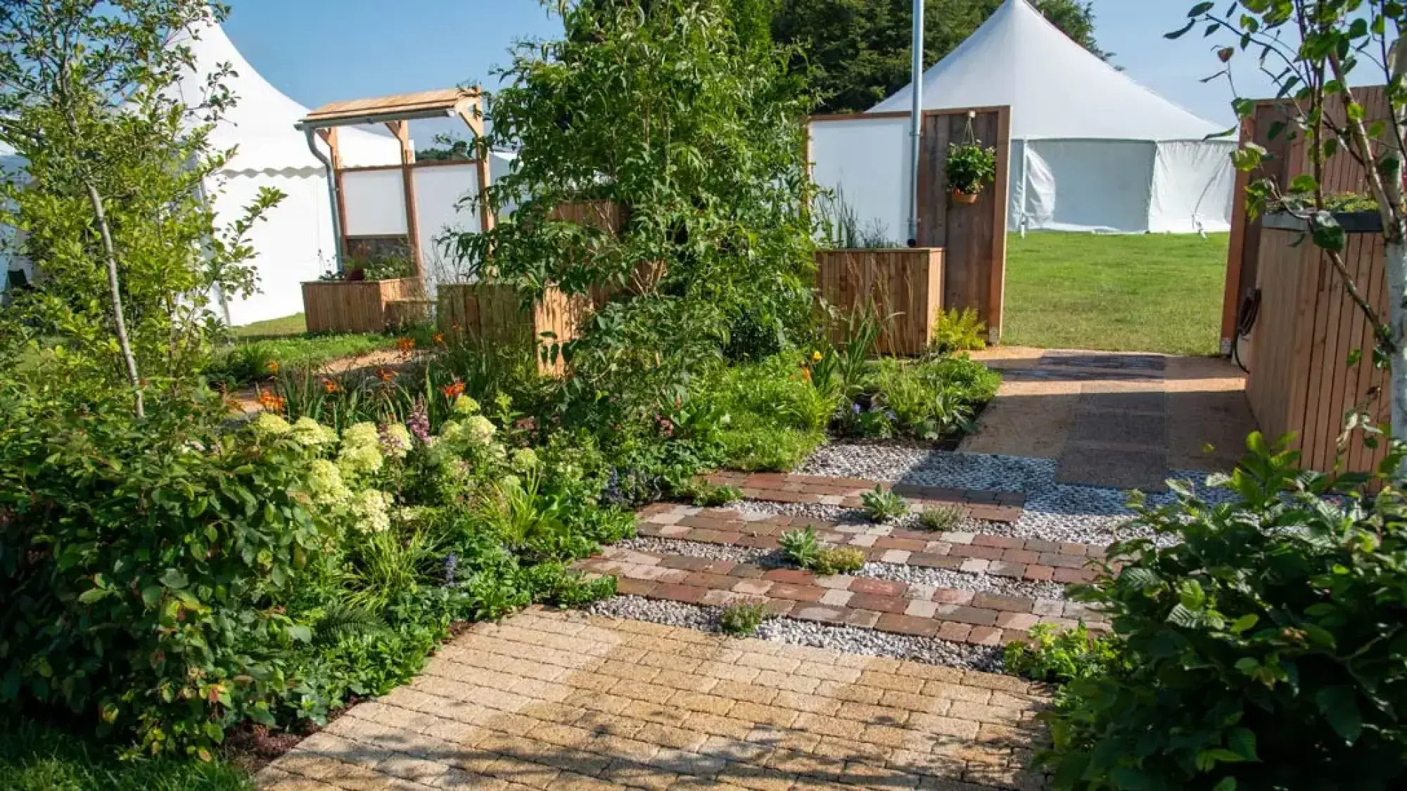 A show garden featuring permeable brick and gravel paths, lush mixed planting, timber raised beds, and a wooden canopy with a rainwater gutter.