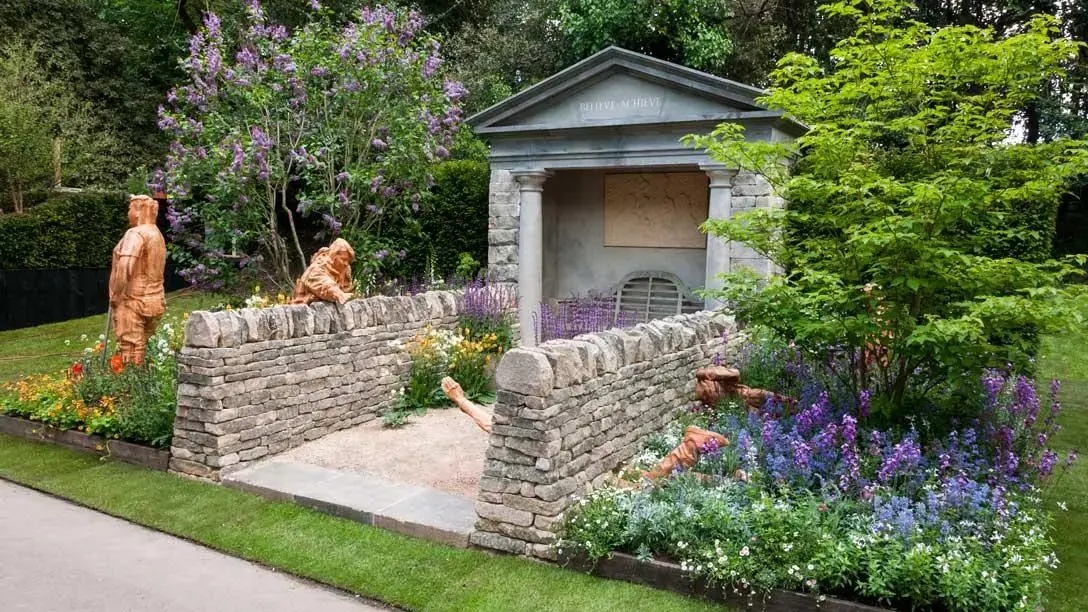An artisan garden features a stone temple, dry stone walls, carved wooden figures, and a paved path winding through lush beds of purple, blue, and white flowers.