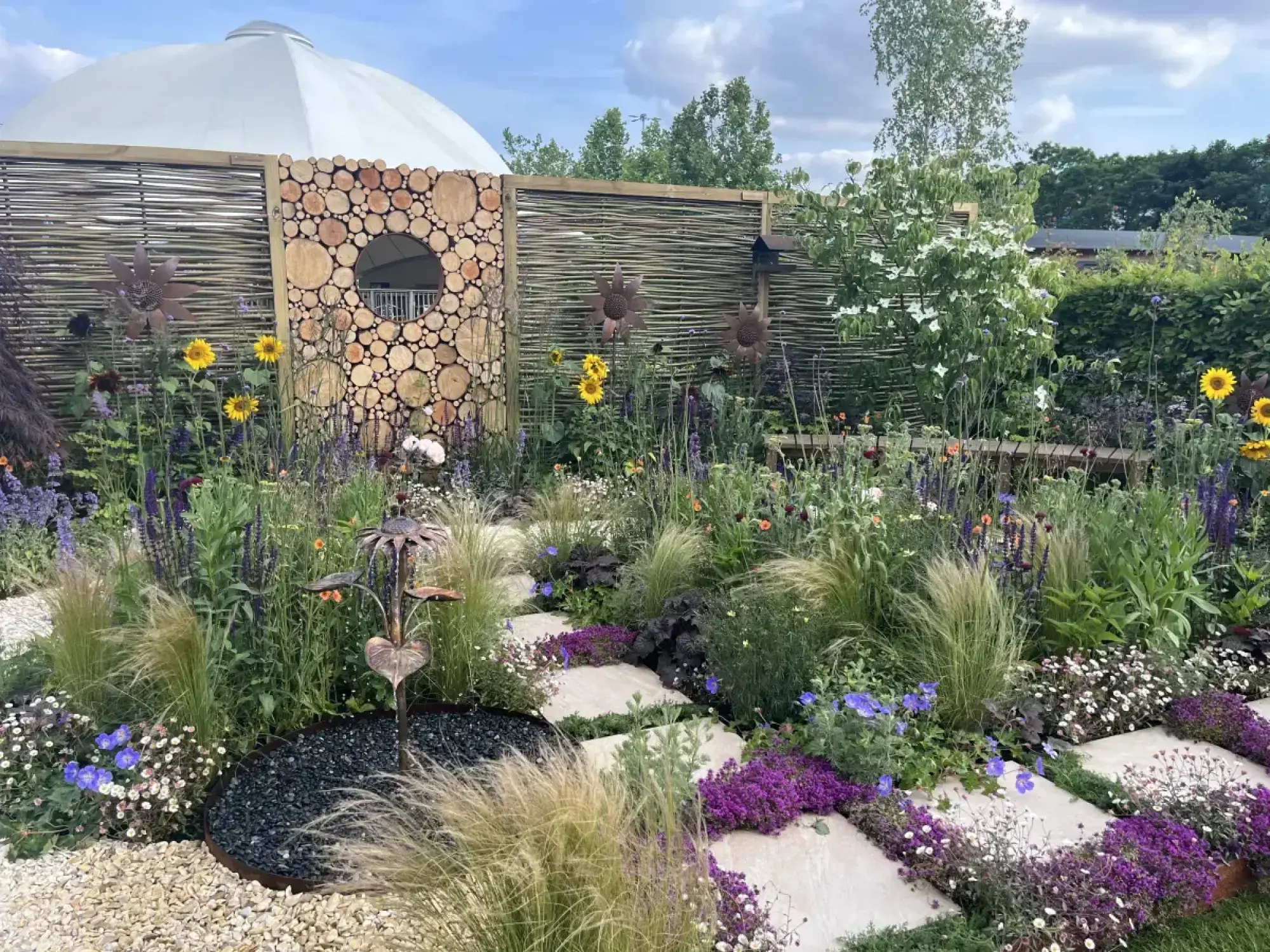 A naturalistic showcase garden with a log round wall, a stone path, yellow sunflowers, ornamental grasses, and mixed vibrant flowers.