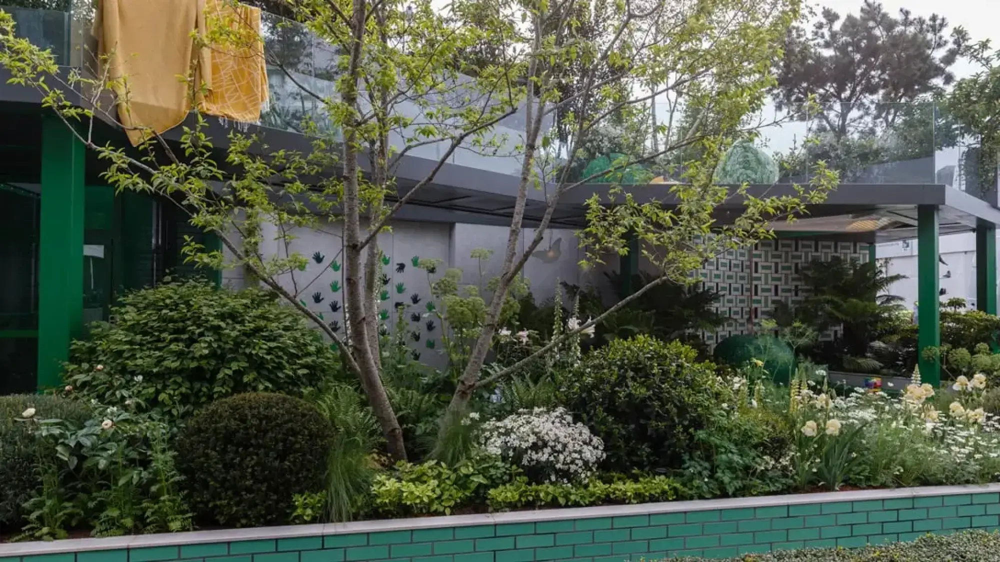 A lush, contemporary show garden features a green brick raised bed, a multi-stemmed tree, white and yellow flowers, and walls with green handprints.