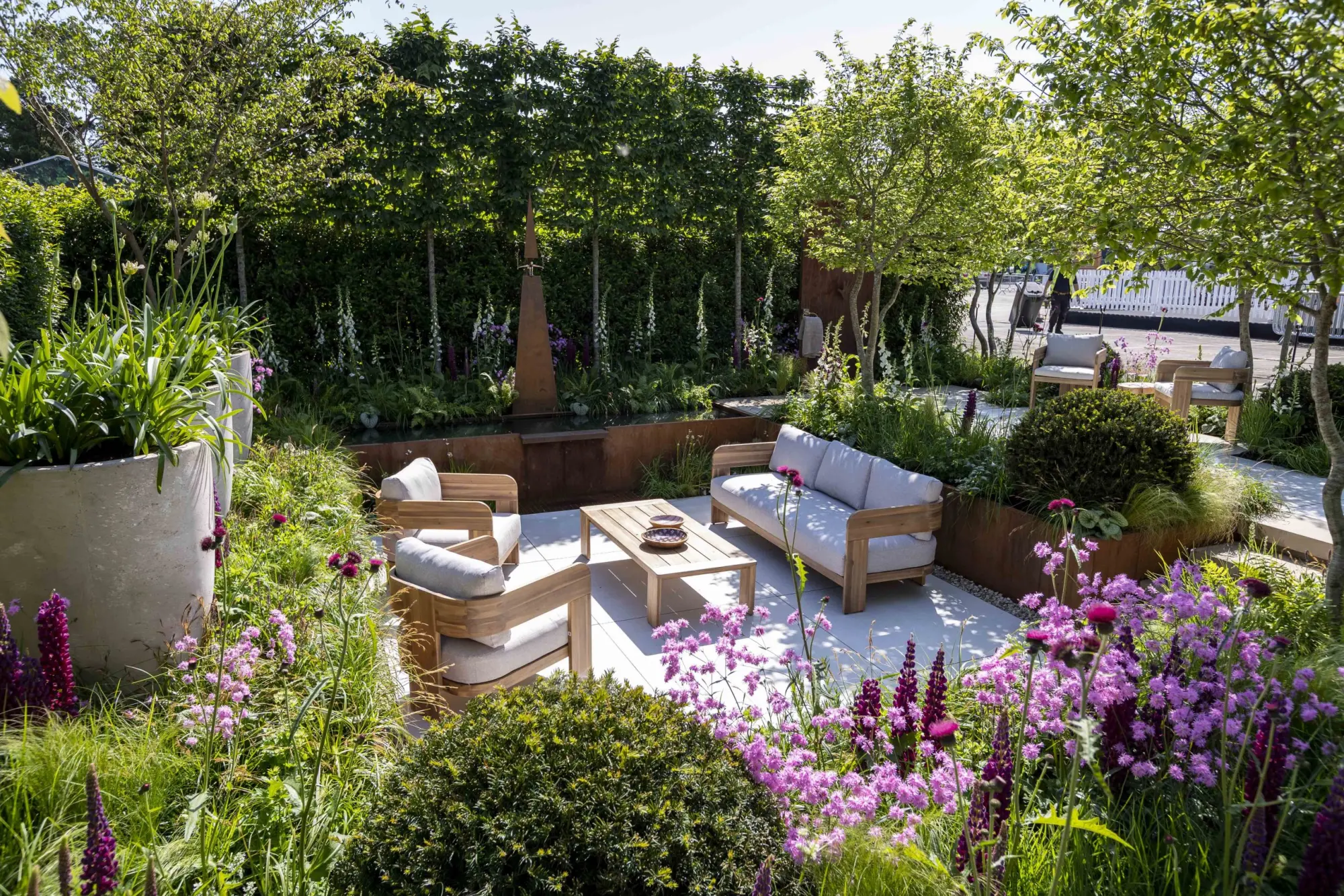 A contemporary garden features a white paved patio with light wood seating, surrounded by lush naturalistic planting in shades of purple and white, a Corten steel water feature, and leafy trees.