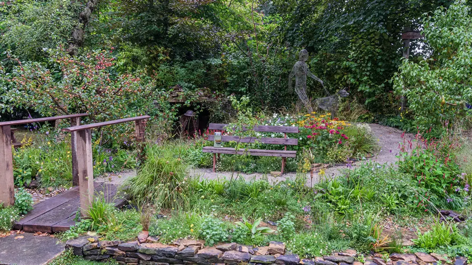 A naturalistic garden features a wire sculpture of a person and guide dog, a rustic wooden bench, and a wooden bridge among wildflowers.