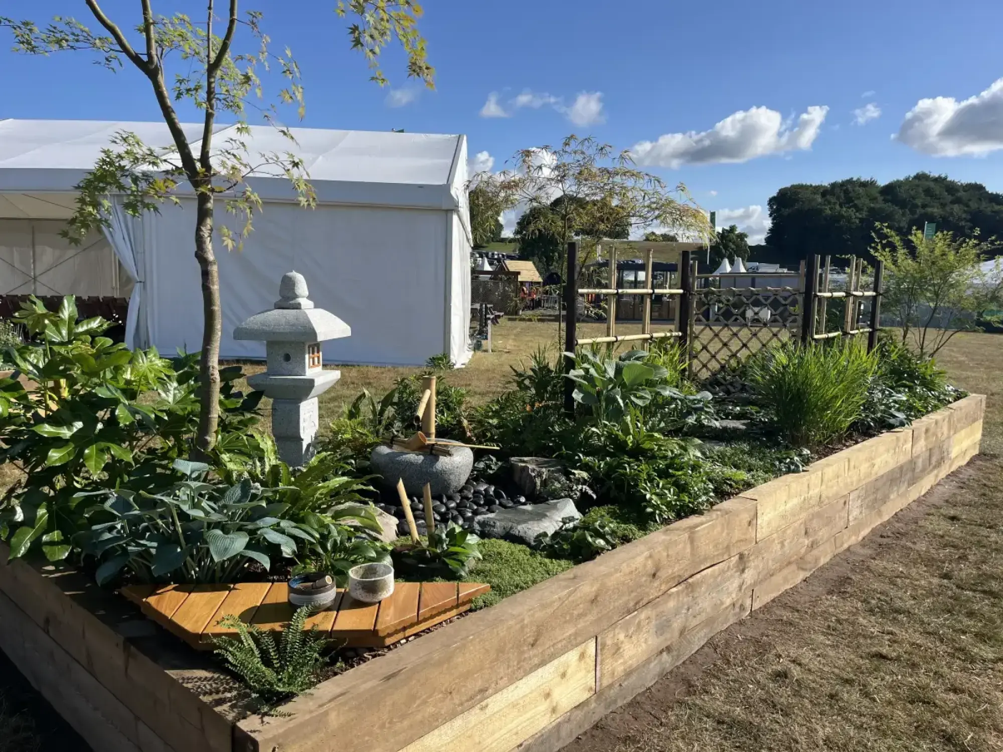 A raised timber garden border features a Japanese style with a stone lantern, bamboo water feature, wooden deck, and lush green hostas, ferns, and an Acer tree.