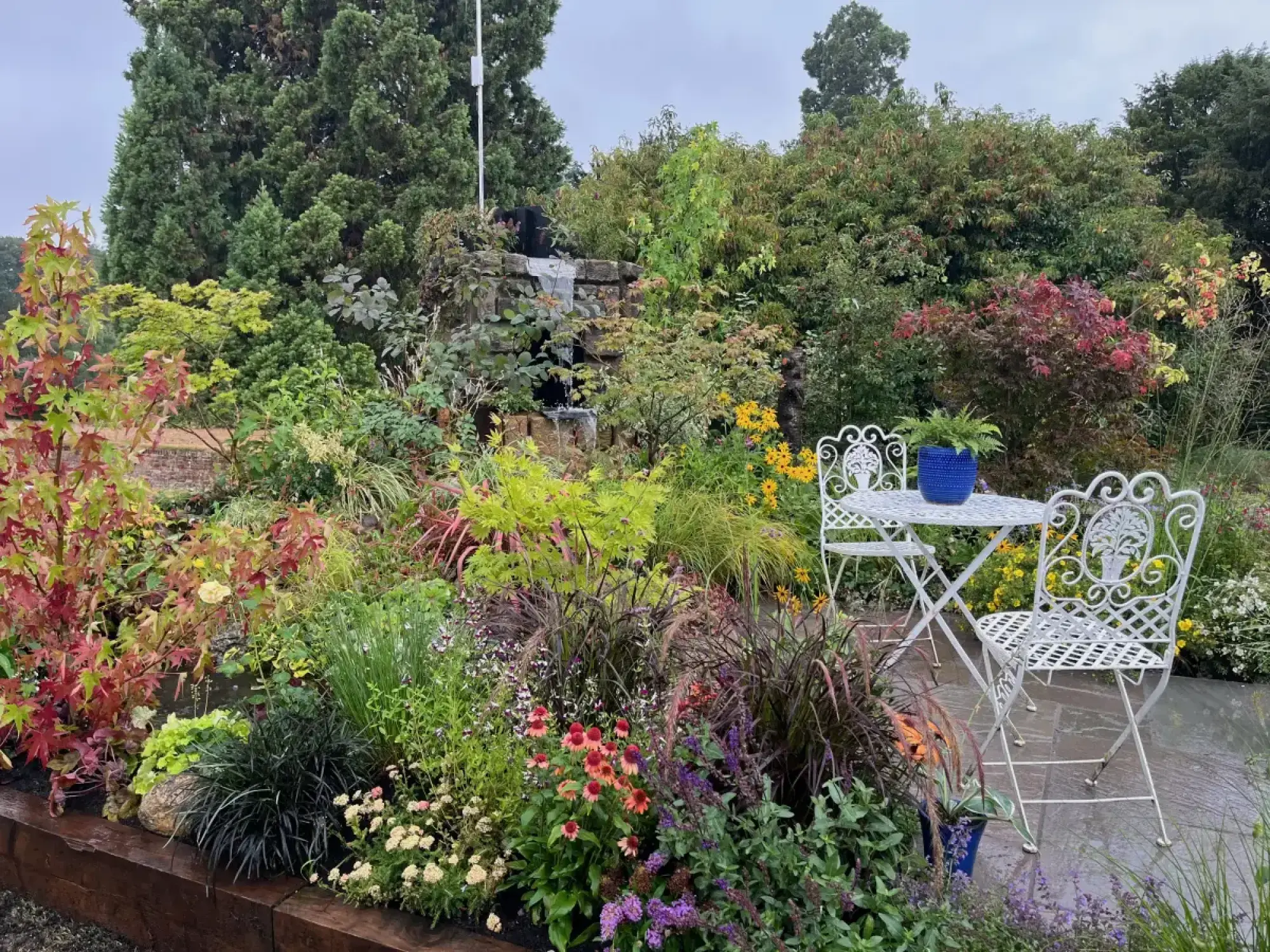 A lush, naturalistic showcase garden features a stone waterfall, vibrant mixed perennials, and a white ornate bistro set on a dark grey patio.
