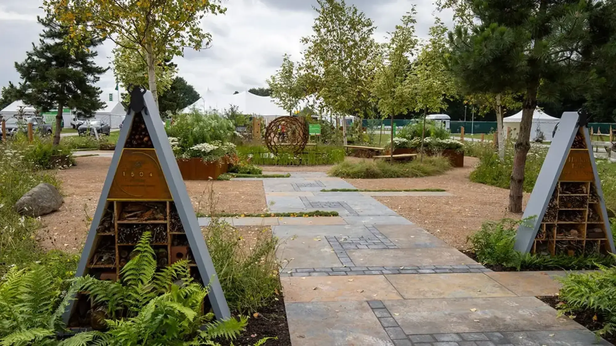 A naturalistic show garden features two tall triangular bug hotels, a rusty metal sphere sculpture, Corten steel planters with integrated benches, and paths winding through wildflowers and grasses.