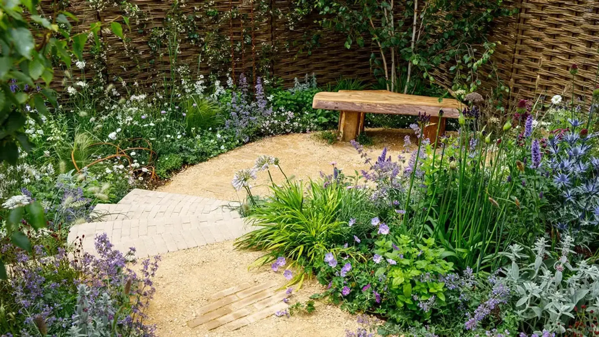 A tranquil Get Started Garden features a curved golden gravel path, a rustic log bench, and abundant informal planting in shades of purple, blue, white, and green, backed by a woven willow fence.