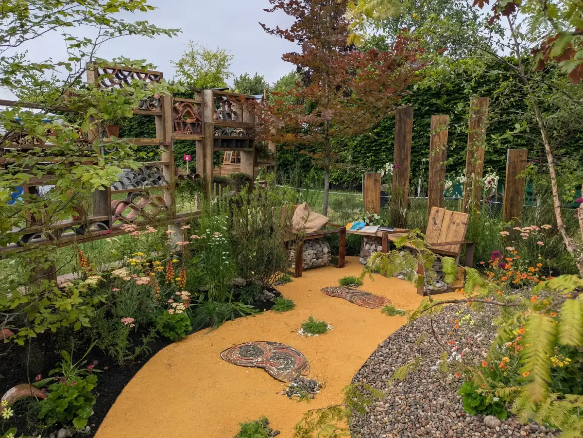A naturalistic show garden featuring a wooden insect habitat wall, gabion seating, and a winding golden pathway with pebble mosaic stepping stones, surrounded by lush wildflower planting.