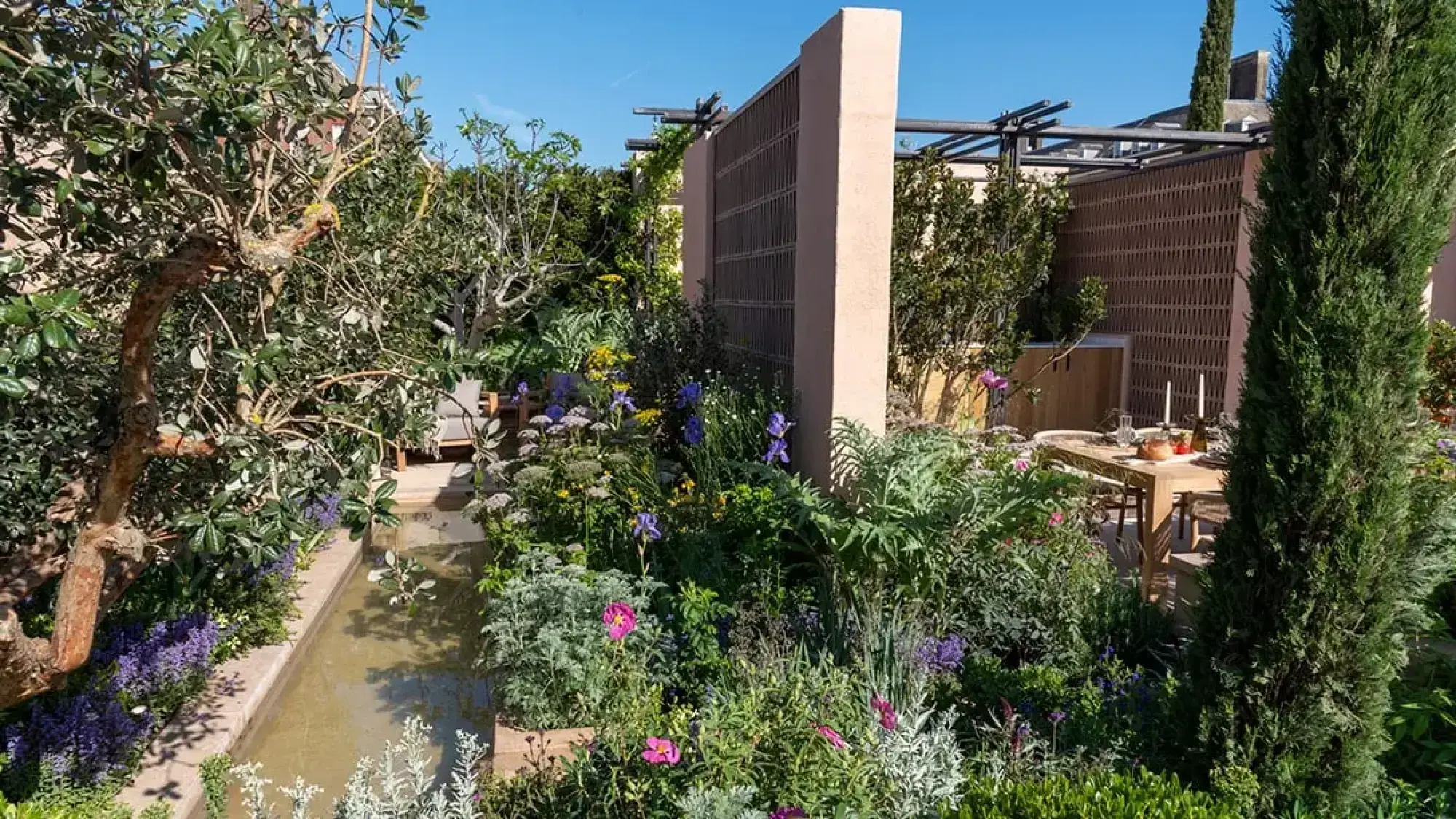 A Mediterranean sanctuary garden with a central water rill, a terracotta wall, outdoor dining, lush planting including blue irises, pink cistus, and tall cypress trees.