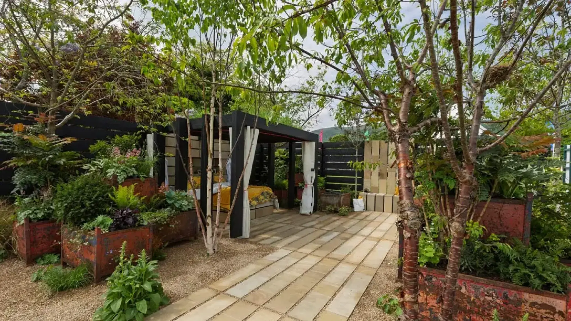 A secluded show garden featuring an outdoor daybed under a black timber pergola, surrounded by lush green foliage in rusted metal planters and a pale paved path.