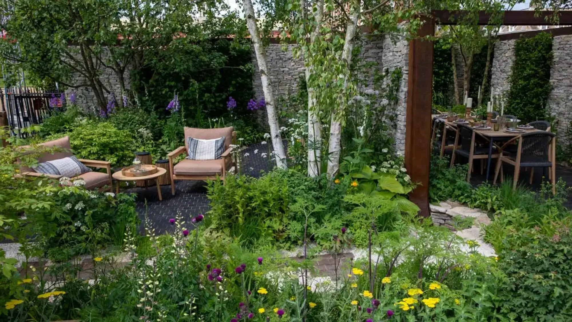 A sanctuary garden with rusted steel pergola, silver birch trees, stone walls, lush planting, and lounge and dining areas.