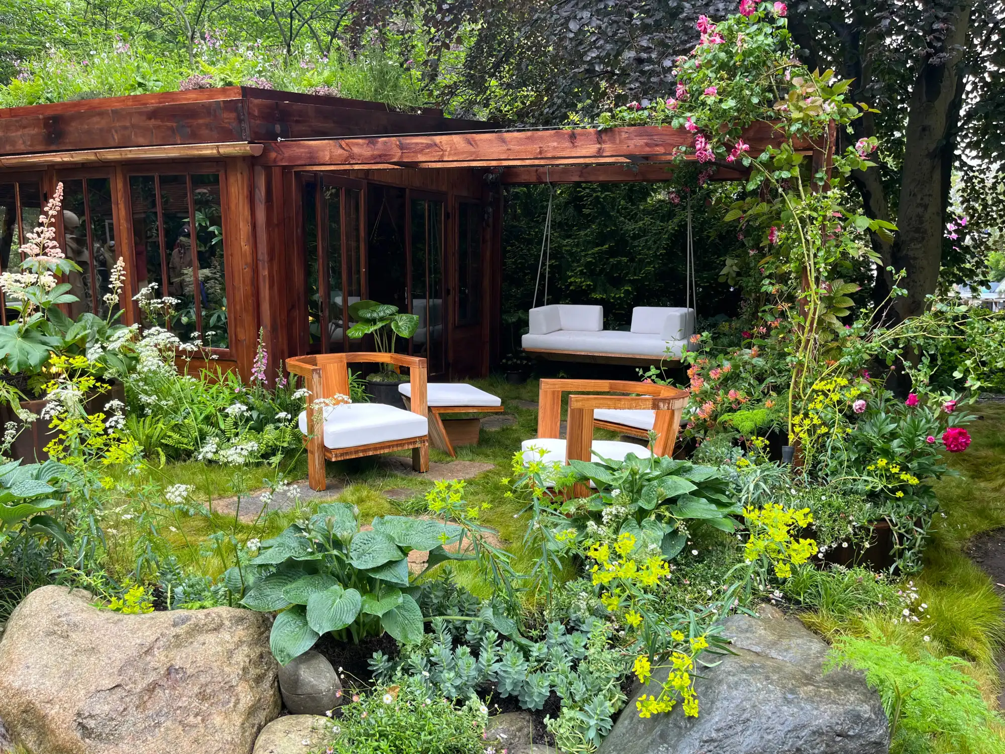 A tranquil garden features a wooden studio cabin, green roof, suspended daybed, wooden seating, lush planting, and natural boulders.