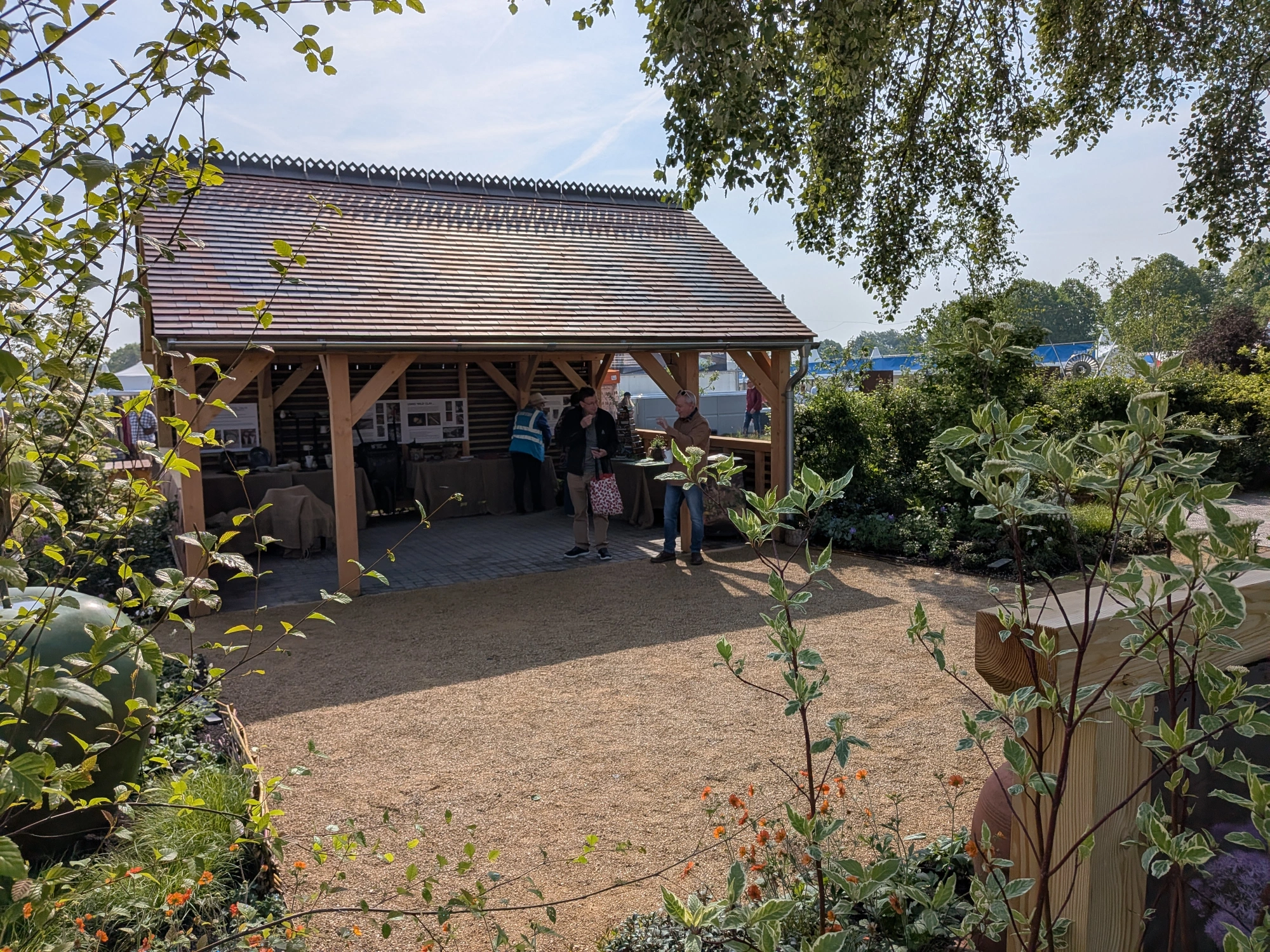 A decorative tiled roof pavilion with a gravel floor is surrounded by mixed planting, featuring orange flowers, variegated shrubs, and a large green pot.
