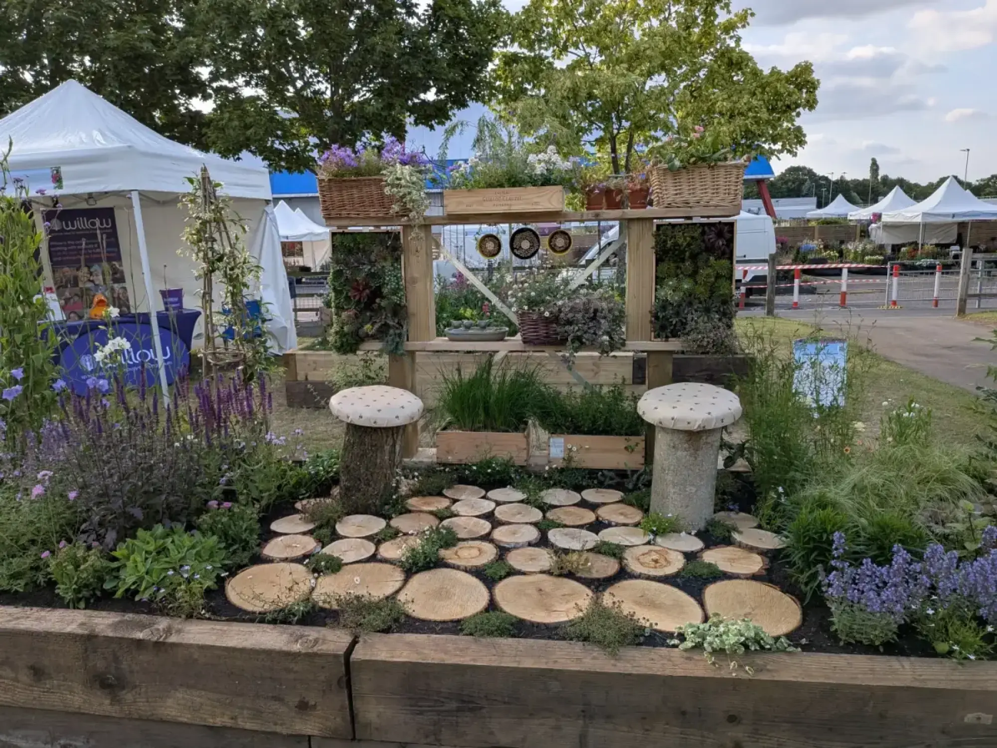 A beautiful border garden features a log slice path, mushroom stools, a wooden structure with vertical planting, and purple and green plants.