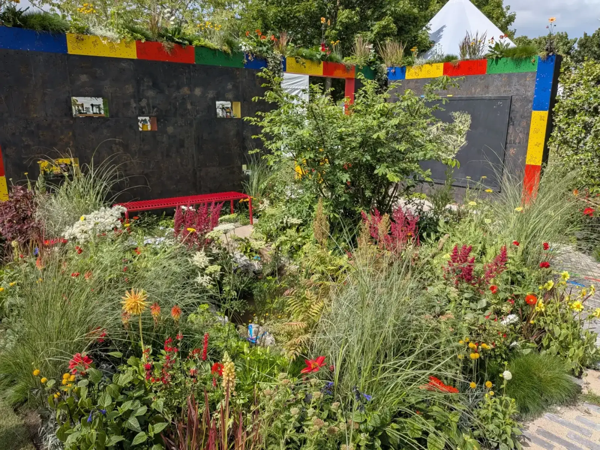 A colourful garden features black walls with colourful LEGO blocks, a red metal bench, and a dense planting of vibrant perennial flowers and tall grasses.