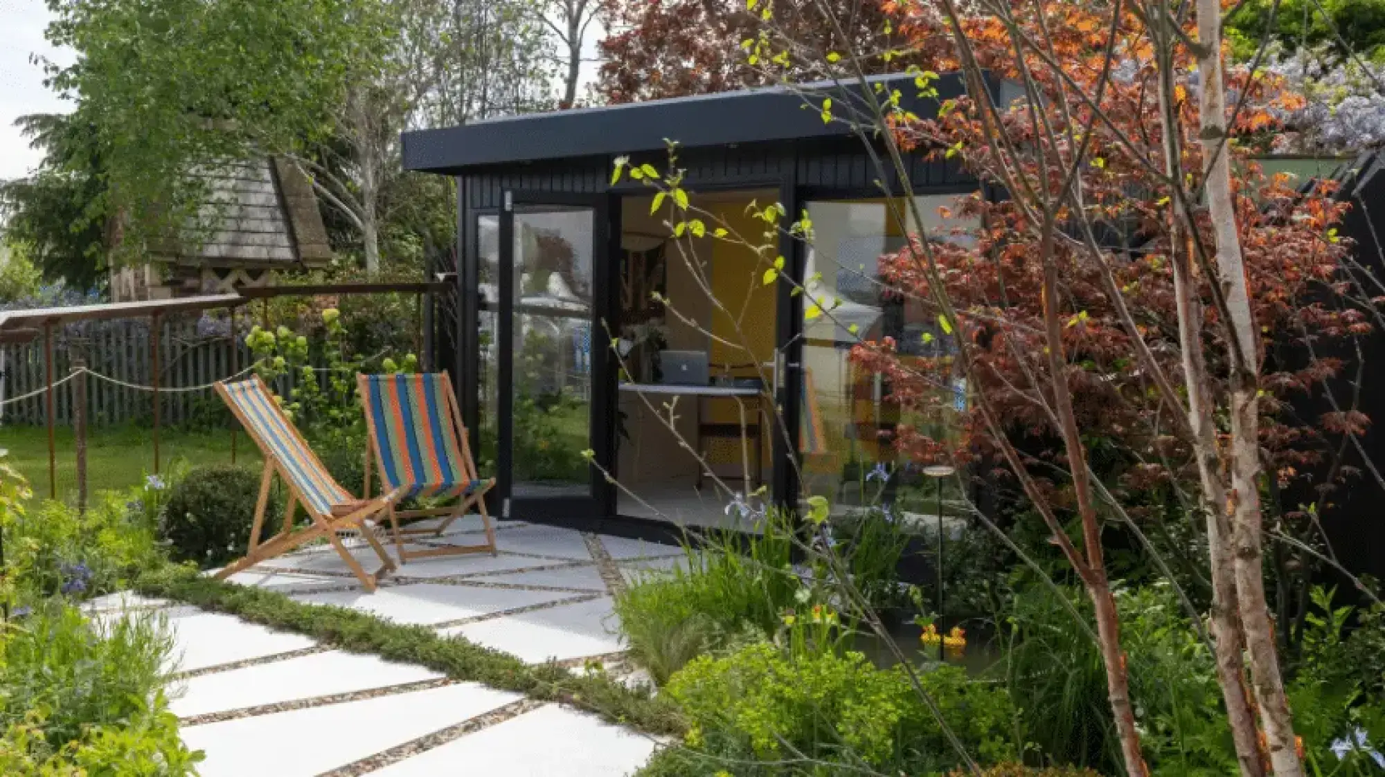 A contemporary show garden with a black modern garden room, white concrete paving, striped deck chairs, birch trees, and a red acer.