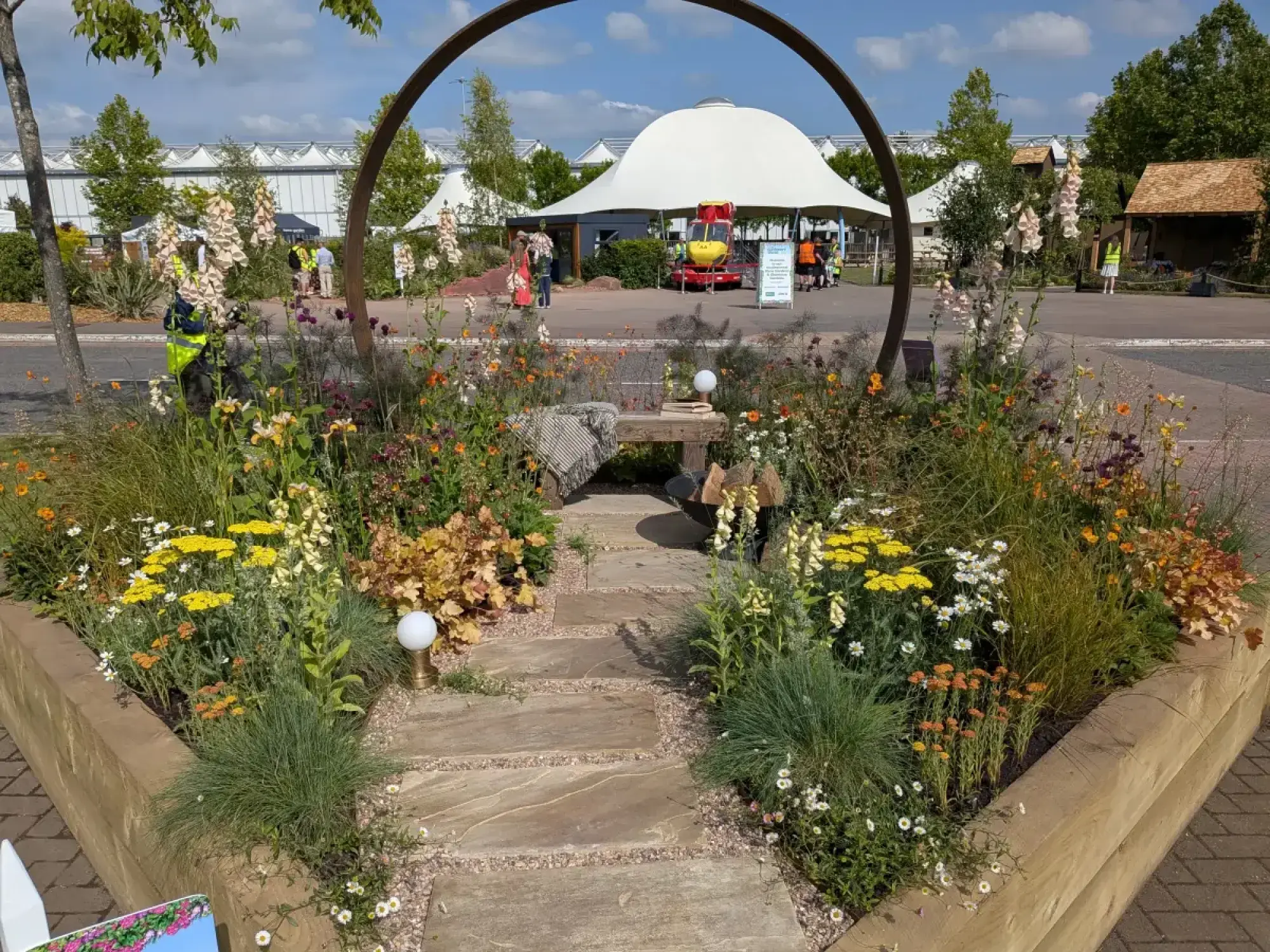 A raised timber garden border frames a natural stone stepping path leading to a wooden bench, all beneath a large circular metal arch, with naturalistic planting of yellow, orange, and cream flowers and grasses.