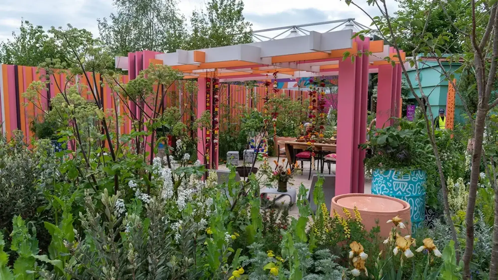 A vibrant feature garden with a pink pergola, slatted walls, hanging flower garlands, a dining area, and lush white and yellow planting.
