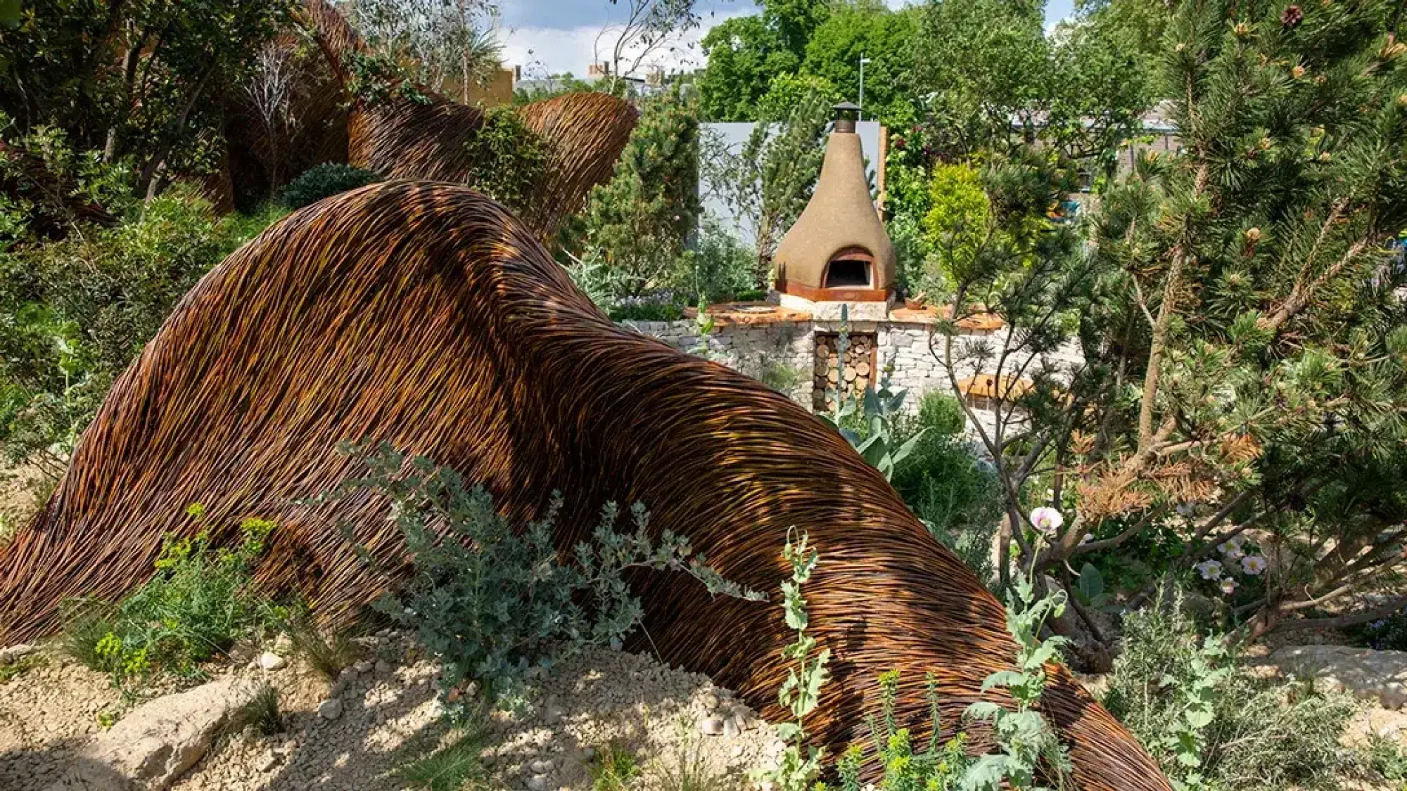 A large, undulating woven willow structure winds through a naturalistic sanctuary garden with a cone-shaped clay oven on a stone wall.