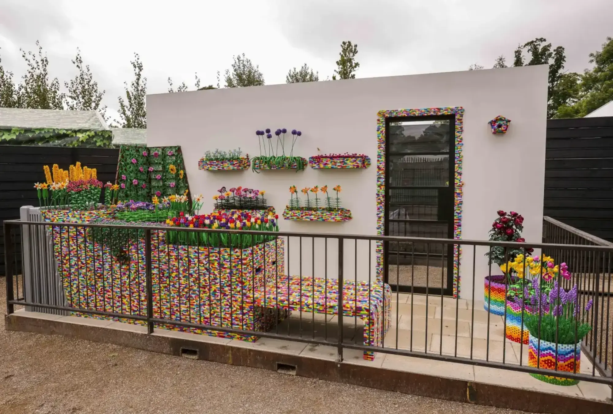 A vibrant balcony garden constructed entirely from multicoloured LEGO bricks, featuring brick flowers in planters, a vertical garden, and a matching bench, set against a white wall.