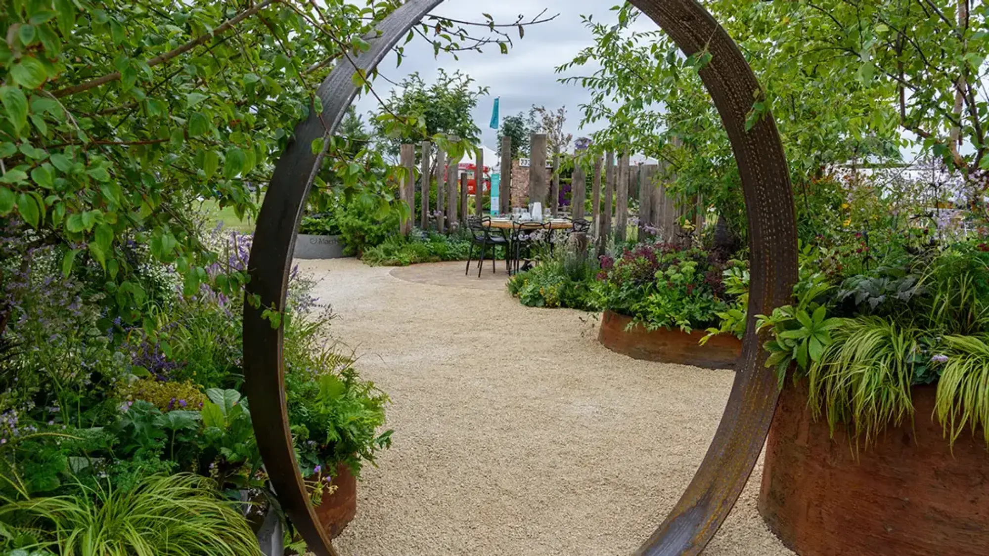 A contemporary feature garden with a large corten steel circular archway leading to a gravel path, a paved dining area with a table and chairs, and lush mixed planting with ferns, grasses, and purple flowers, framed by a timber log screen.