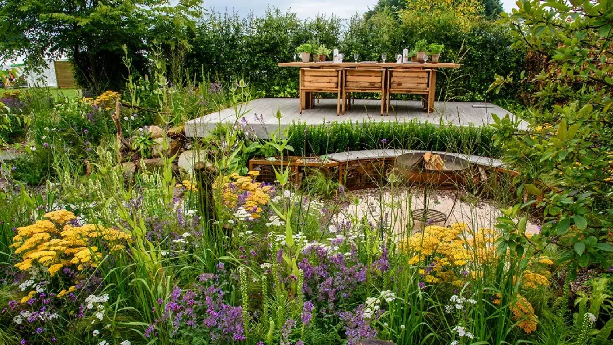Grey deck with rustic table overlooks gravel fire pit, surrounded by yellow and purple meadow planting.