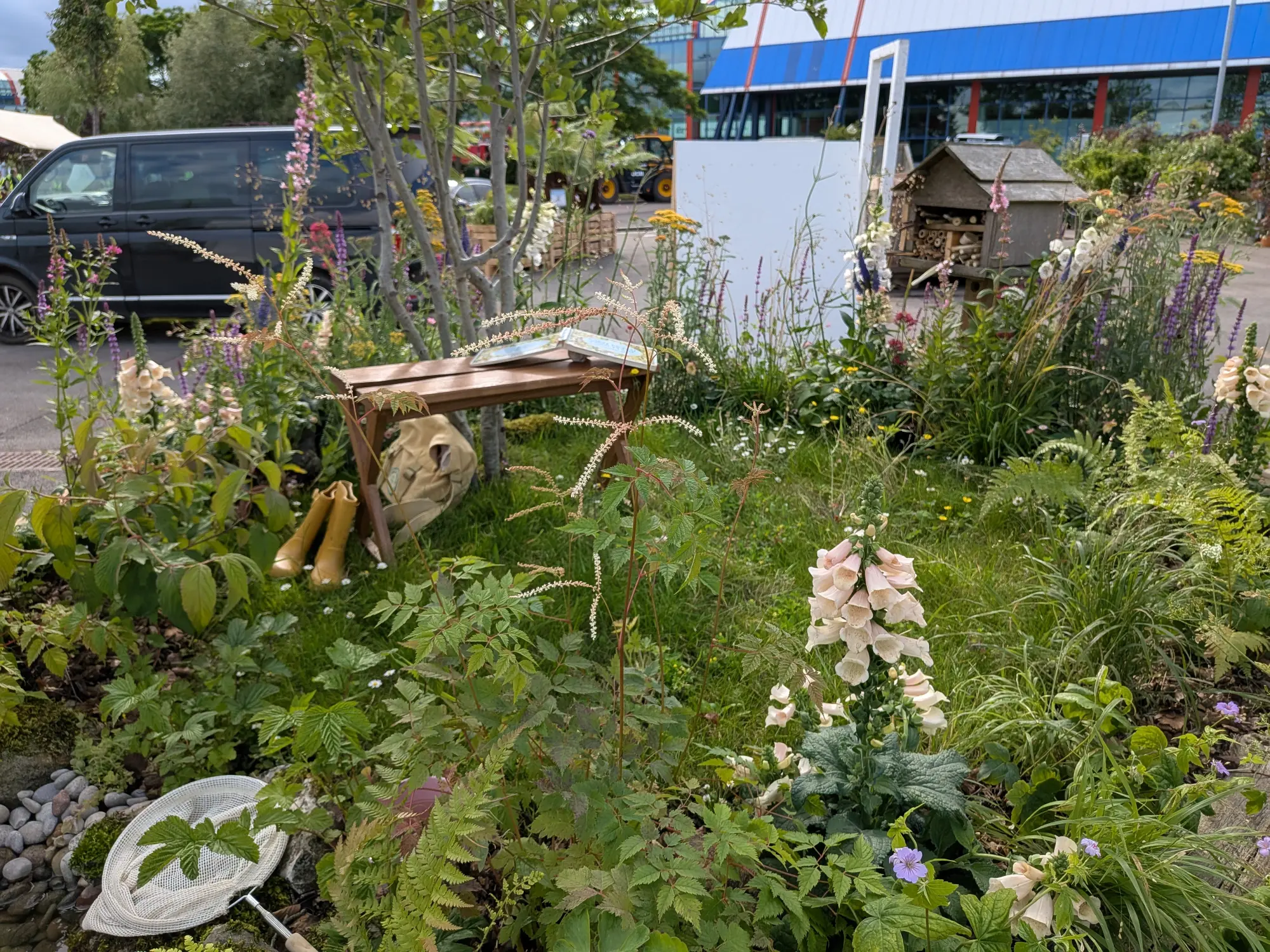 An informal garden border features a wooden bench with open books, yellow wellington boots, a butterfly net, and an insect hotel, surrounded by lush naturalistic planting of pale pink foxgloves, purple spires, white feathery flowers, and ferns.