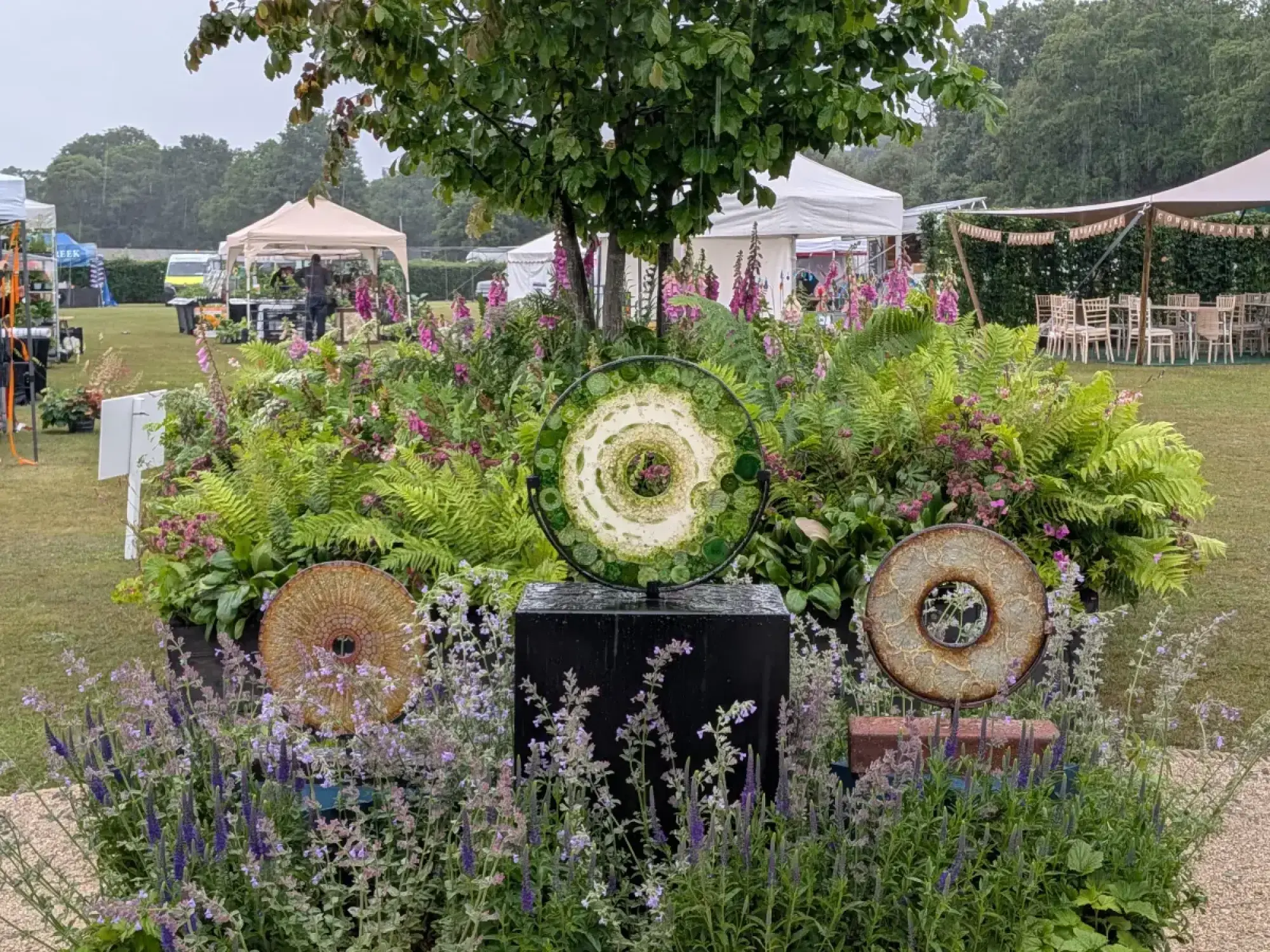 A show garden features a green and white spiral sculpture on a black pedestal, flanked by rusted rings, surrounded by ferns, foxgloves, and salvia.