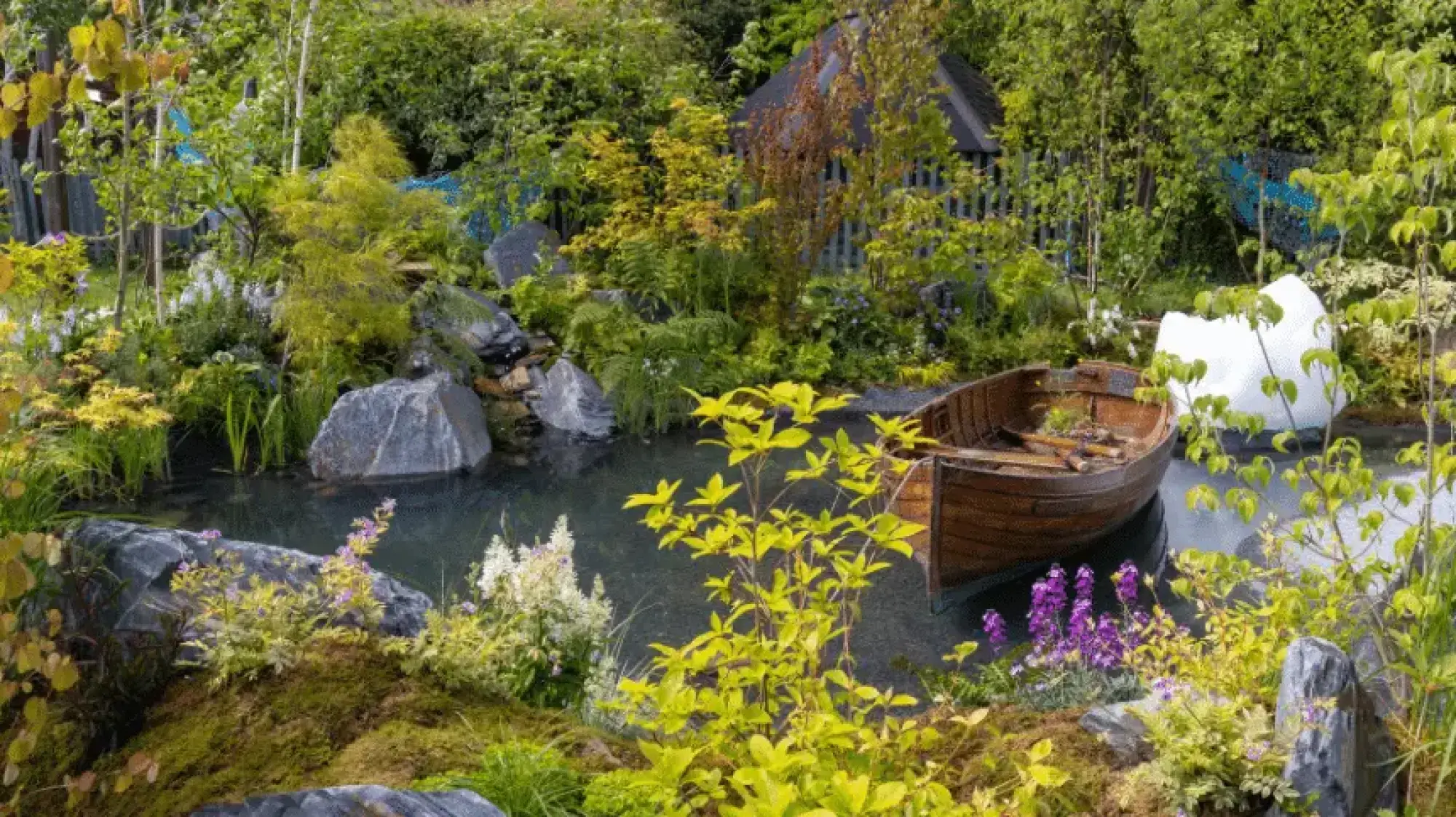 A wooden rowboat rests in a dark pond surrounded by lush greenery, large grey boulders, and a distinctive white iceberg-shaped sculpture.
