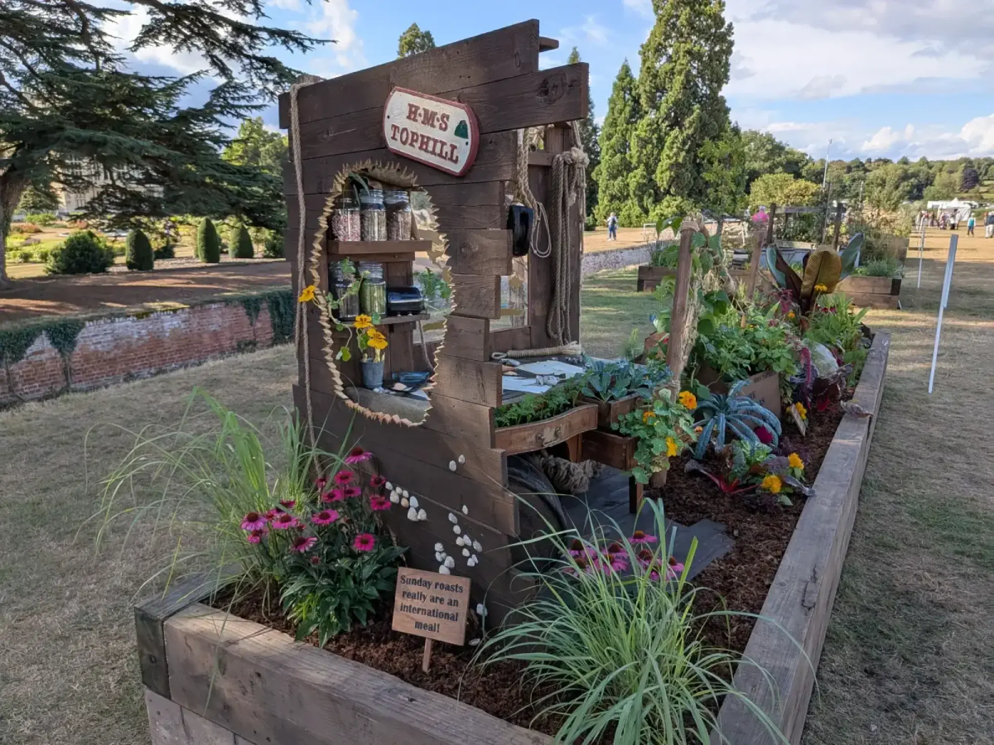 A rustic raised border garden features a wooden ship hull structure with an 'H-M-S Tophill' sign, jars, and chard, kale, and coneflowers.