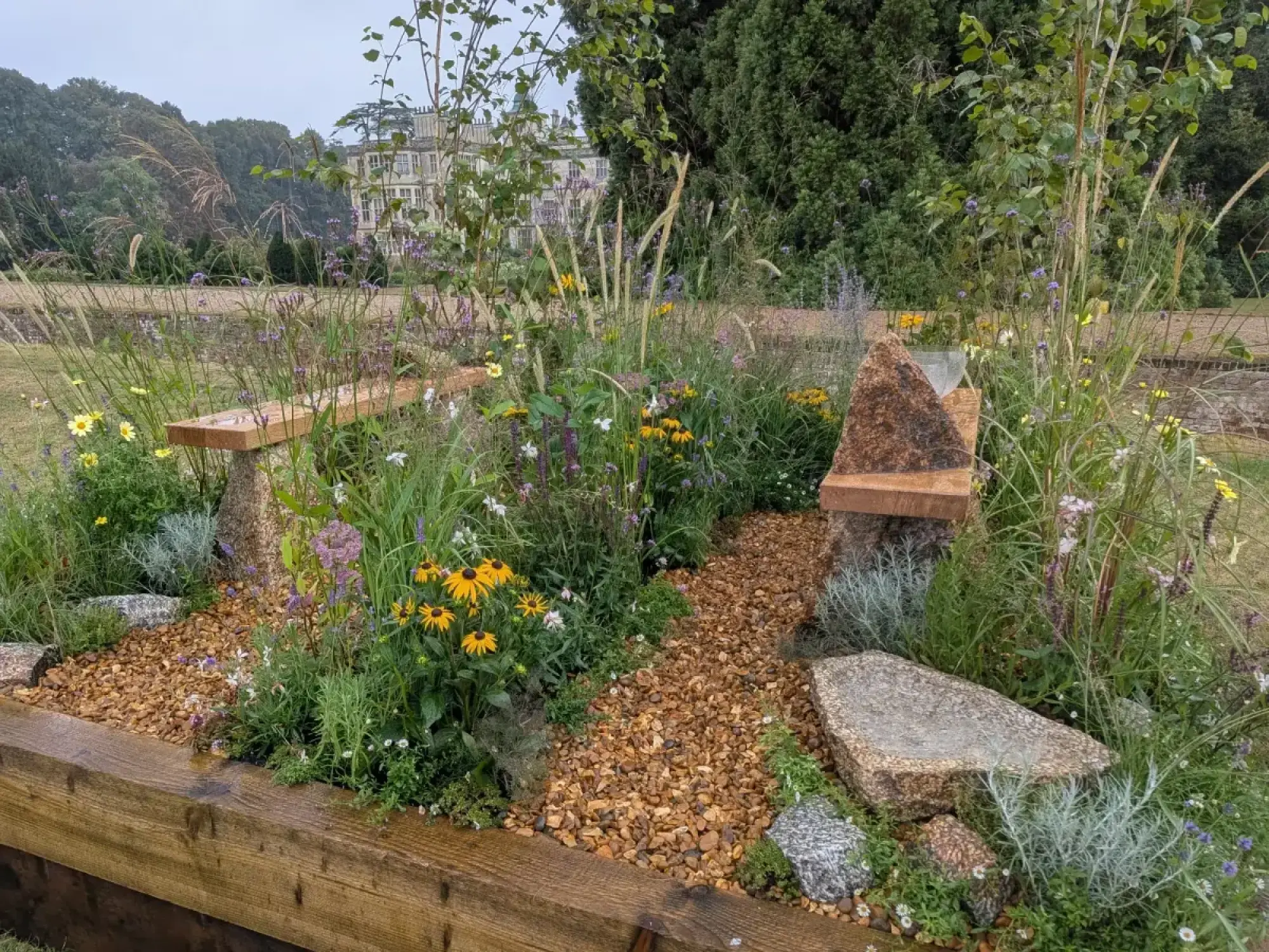 A naturalistic border garden has a pebble path, wooden benches on stone, a triangular water feature, and meadow planting with yellow and purple flowers.