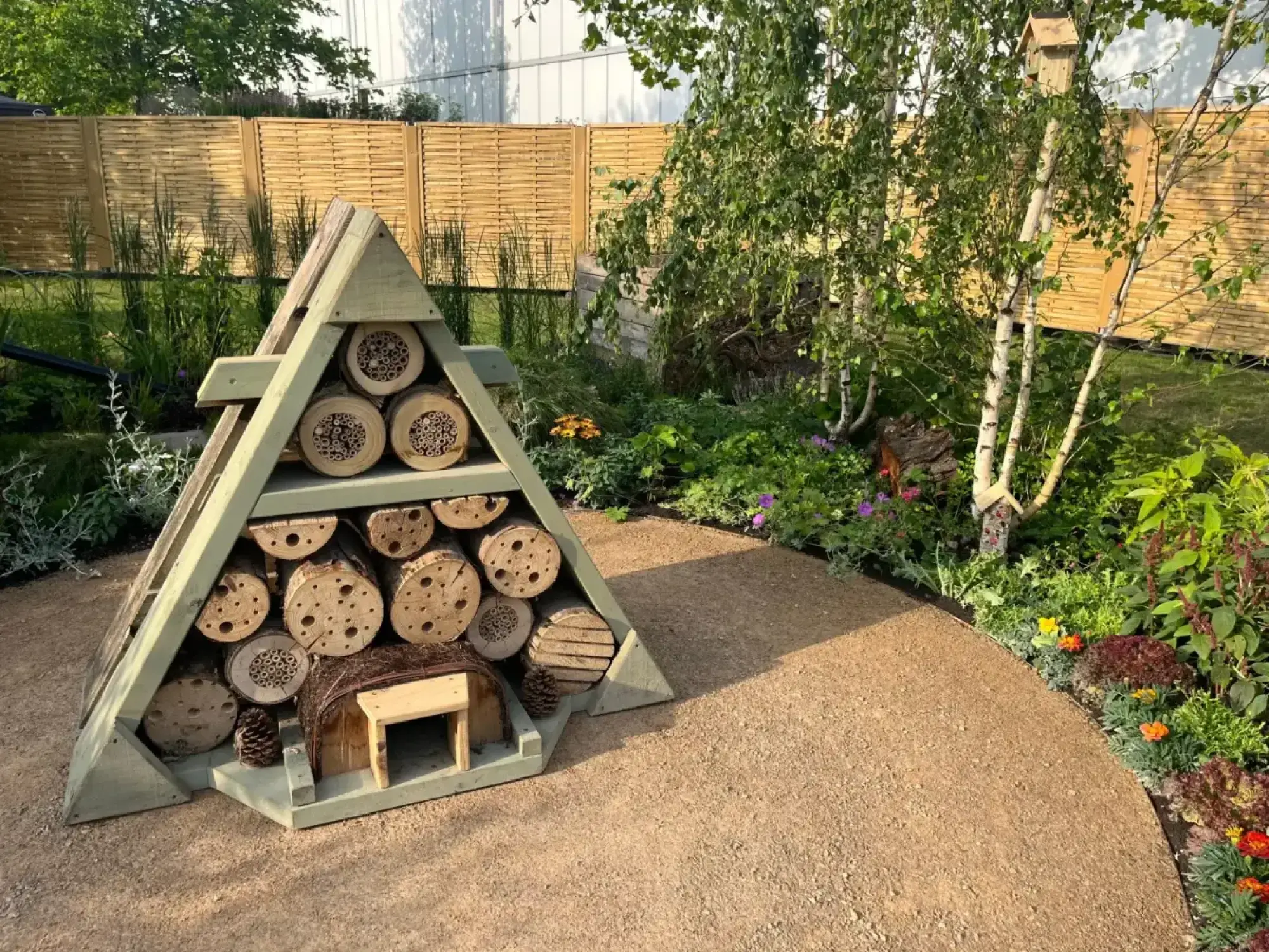 A pyramid bug hotel with drilled logs is on a gravel path, bordered by naturalistic planting, white birch trees, and a wooden birdhouse.