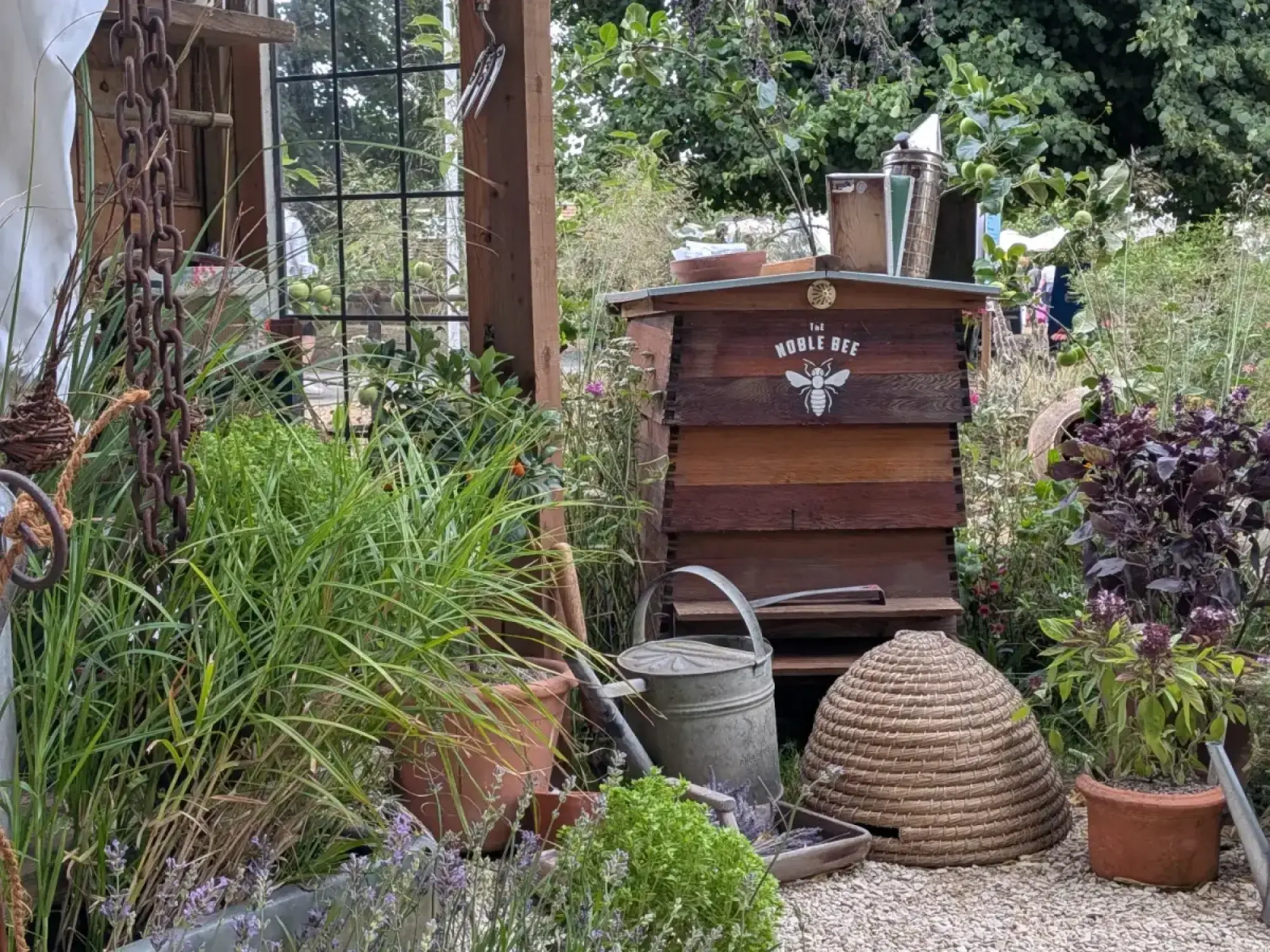 A rustic garden features a wooden beehive, a traditional woven skep, a galvanized watering can, and lush plantings of lavender, grasses, and purple basil.