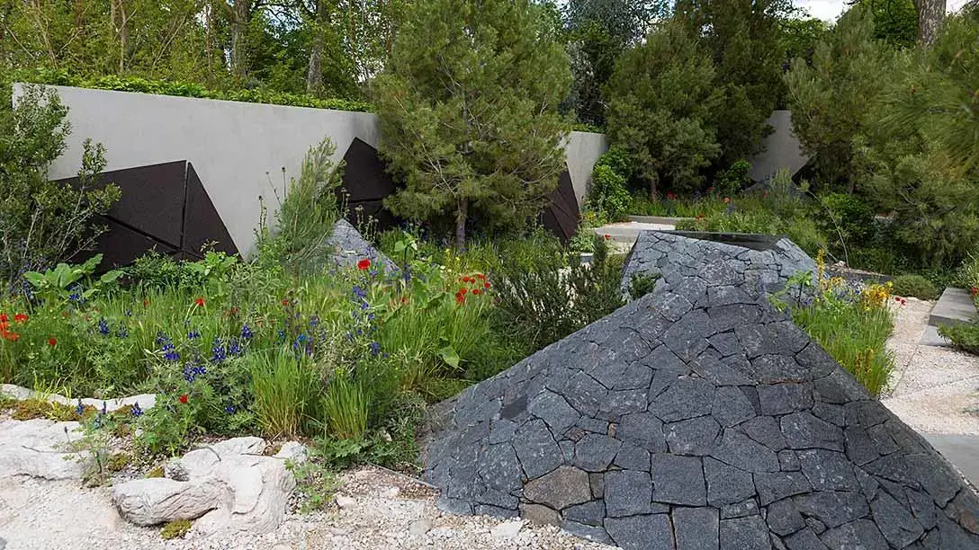 A contemporary show garden features dark, angular dry-stacked stone structures, a grey concrete wall with dark geometric panels, and naturalistic planting with red poppies and blue delphiniums.