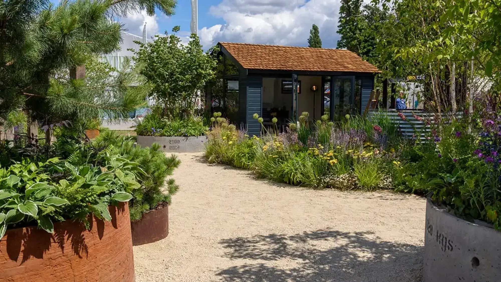 A dark timber cabin with a shingled roof is visible beyond a winding gravel path, bordered by vibrant perennial plants and large planters.