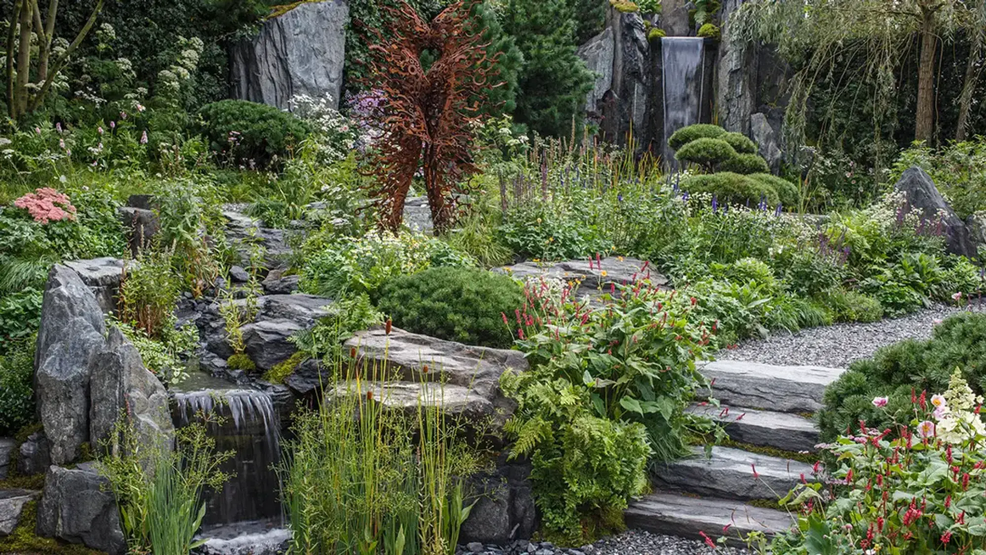 A rugged show garden features dark slate rocks, cascading waterfalls, a rusted tree sculpture, and lush green plants with colourful flowers.