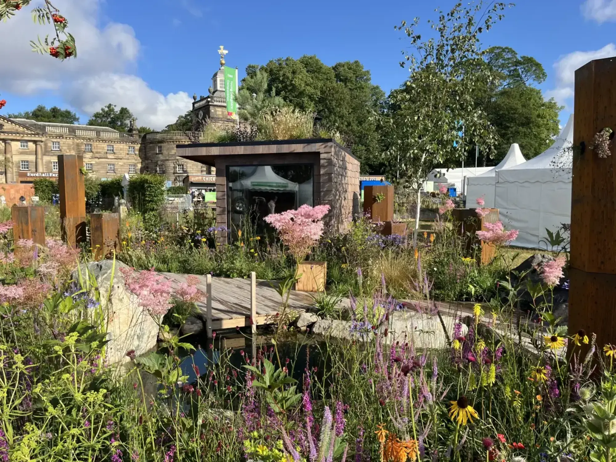 A naturalistic garden features a wooden shed with a green roof, a pond, jetty, and rusty metal posts among colourful wild perennials.
