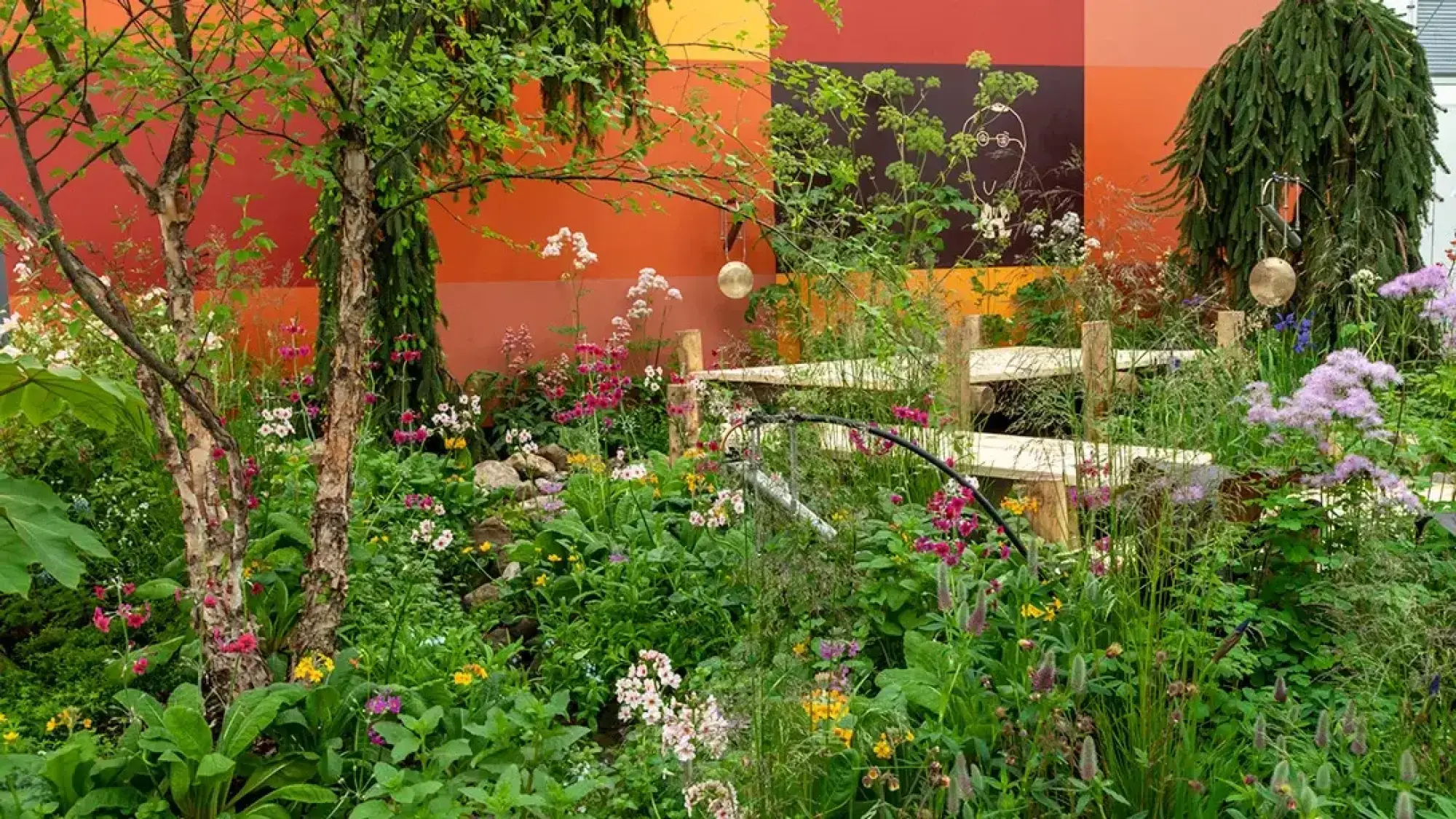 A lush, naturalistic garden with wooden steps, suspended gongs, and vibrant pink, white, yellow, and purple flowers against a warm-toned mural.
