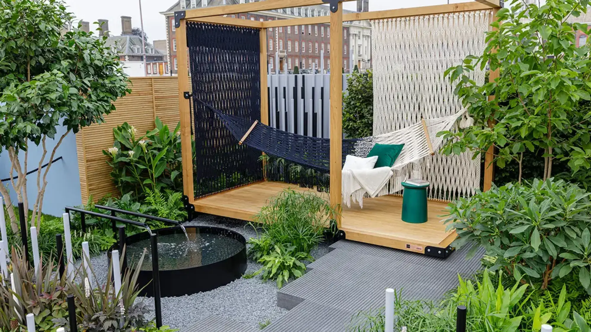 A modern sanctuary garden featuring a wooden structure with black and white macrame screens, two hammocks, a raised deck, a circular water feature, and lush green foliage.