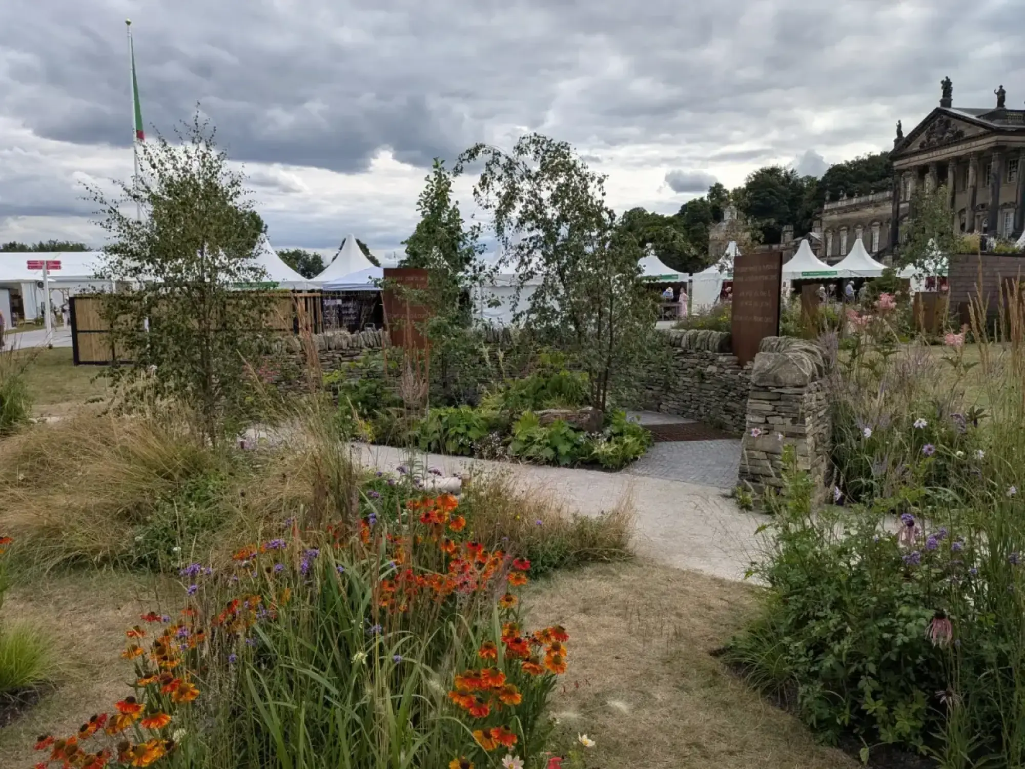 A naturalistic garden features dry stone walls, a light-coloured path, and beds filled with ornamental grasses and orange, purple, and pink wildflowers.