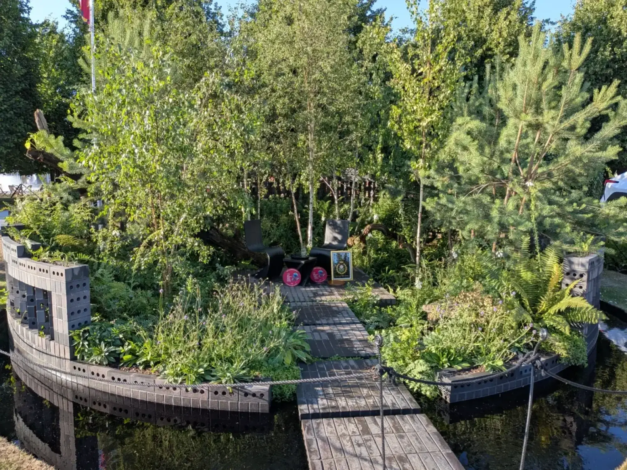 A tranquil, modern island garden called "The Subaru Cocoon," featuring a boat-shaped planter made of dark grey blocks, lush trees and ferns, and a wooden walkway leading to two black chairs.