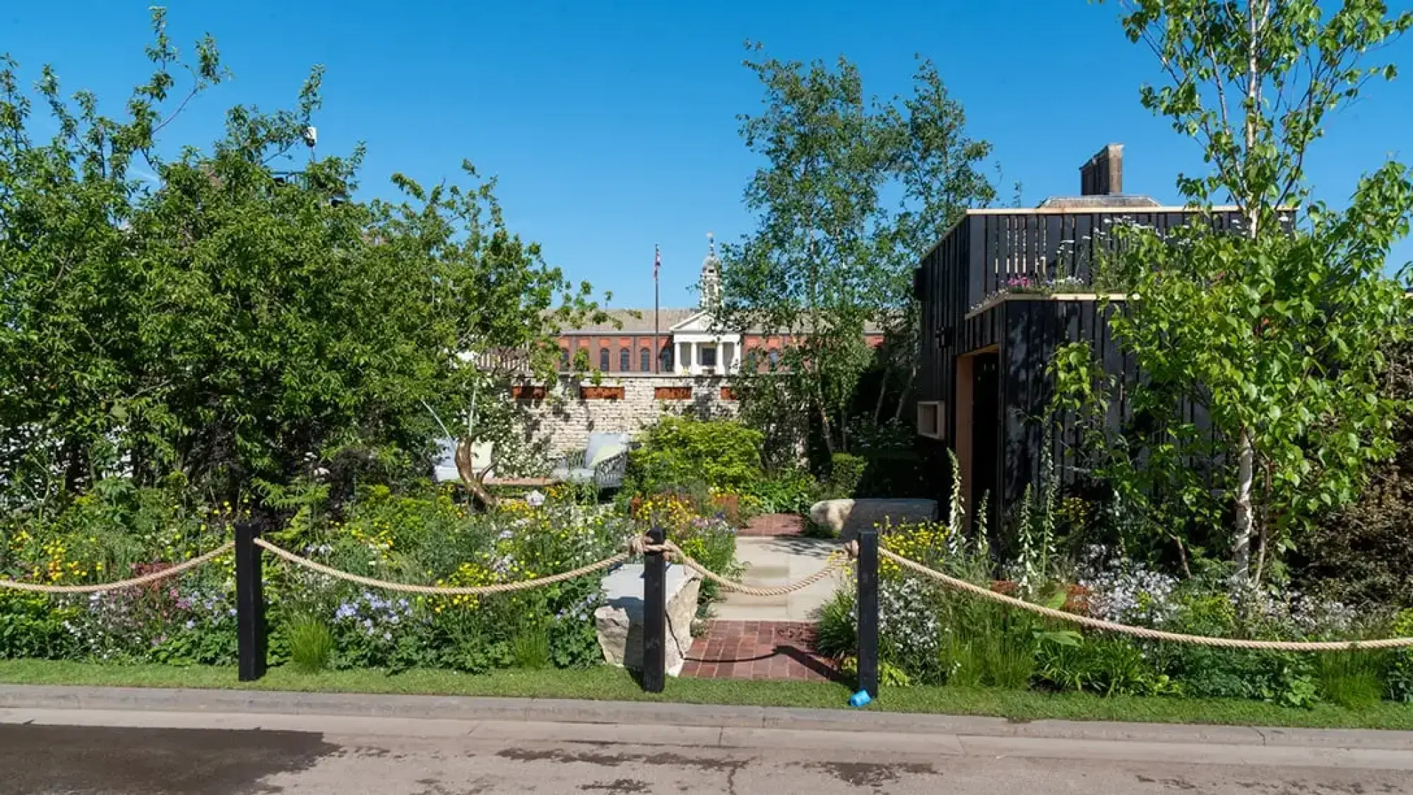A tranquil sanctuary garden with a dark timber cabin, green roof, curved brick path, natural boulders, and abundant wildflowers and trees.